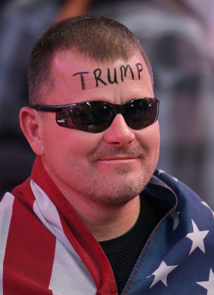 Man wearing sunglasses, draped in an American flag, with "TRUMP" written on his forehead