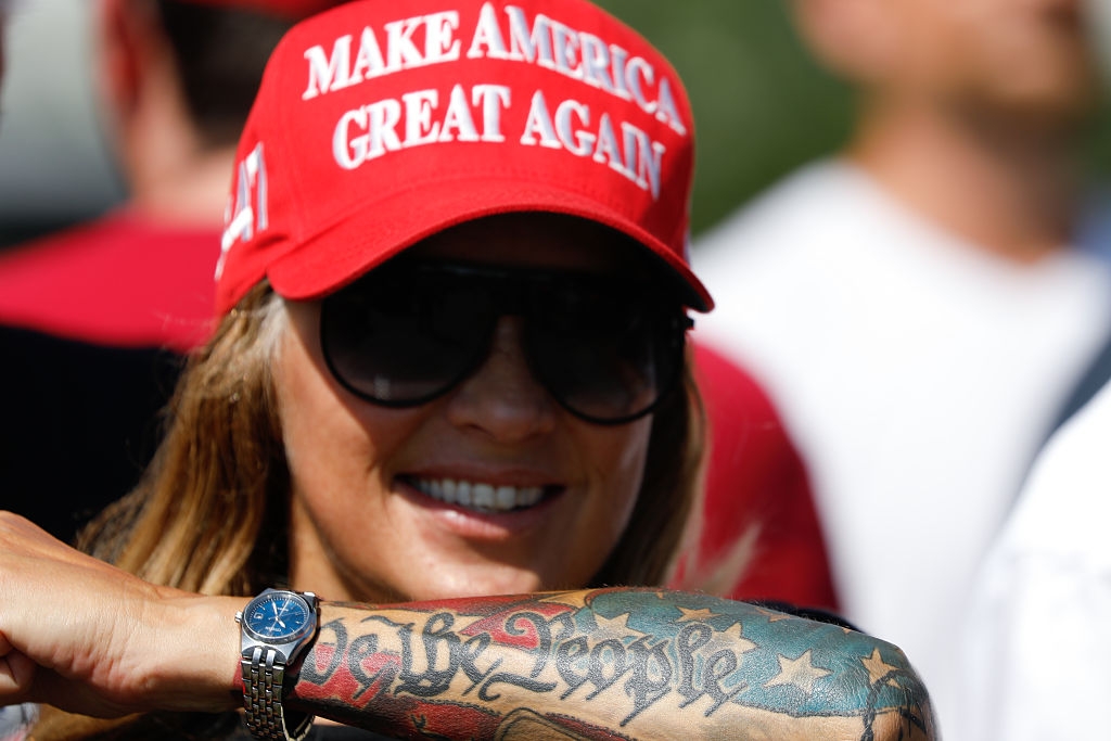 Person som har på seg en "Make America Great Again" hatt og solbriller, smilende og viser en tatovering med dekorativ tekst på armen