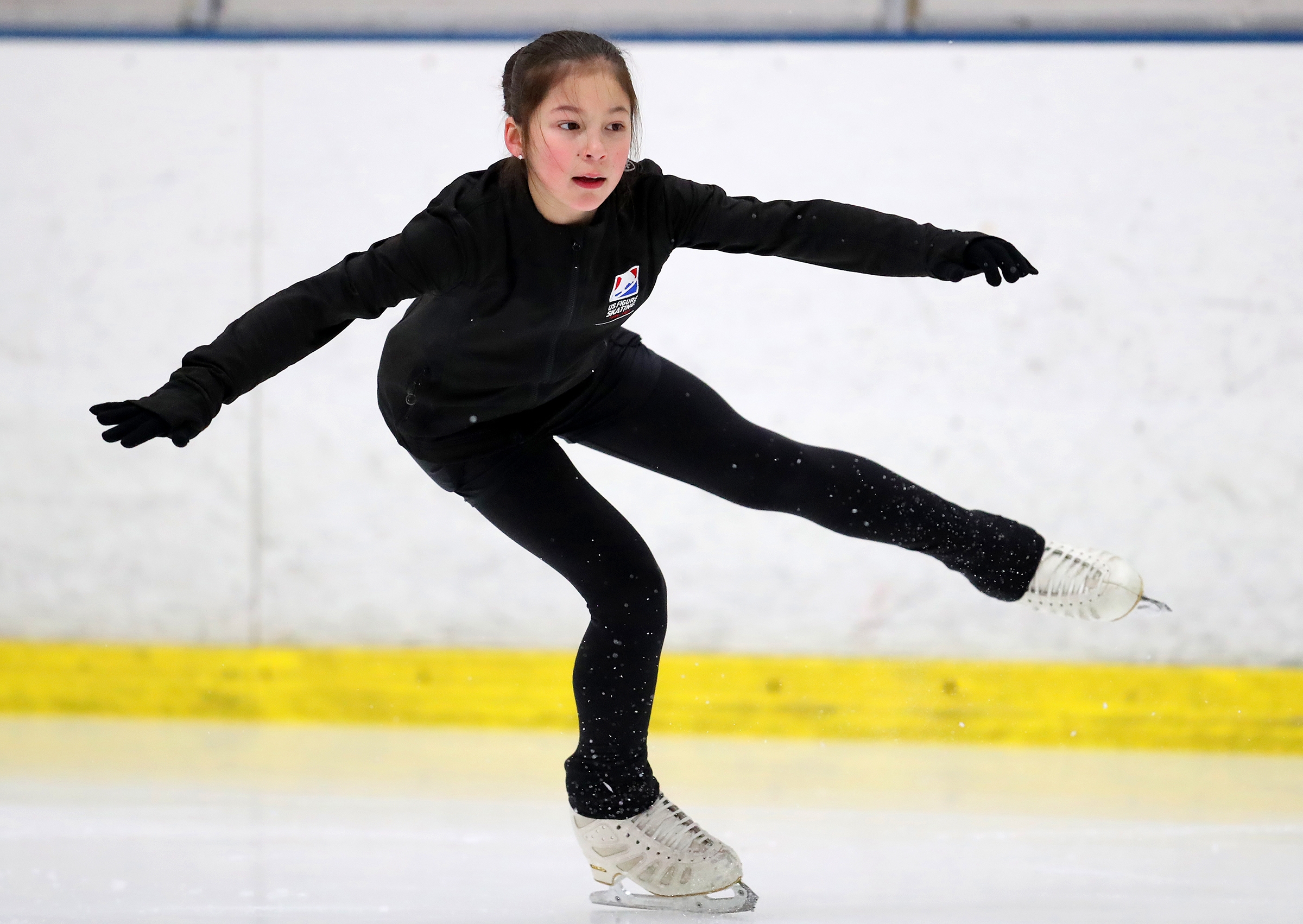 En kunstløper opptrer på is, balanserer på ett ben med fokusert uttrykk, iført et elegant svart antrekk og skøyter