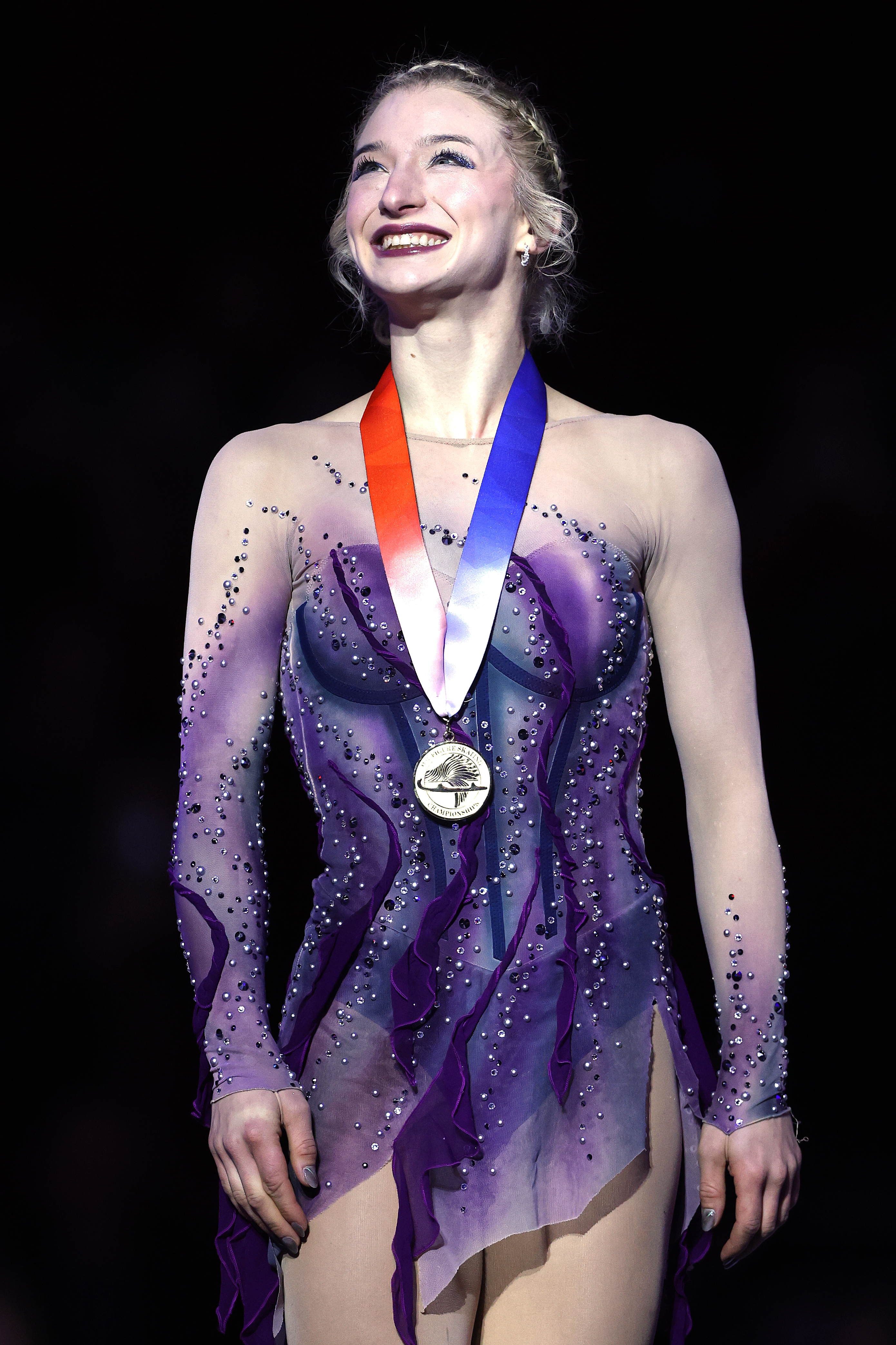 En kunstløper smiler bredt, iført en glitrende, ren kjole og en medalje rundt halsen, stående på en mørk bakgrunn