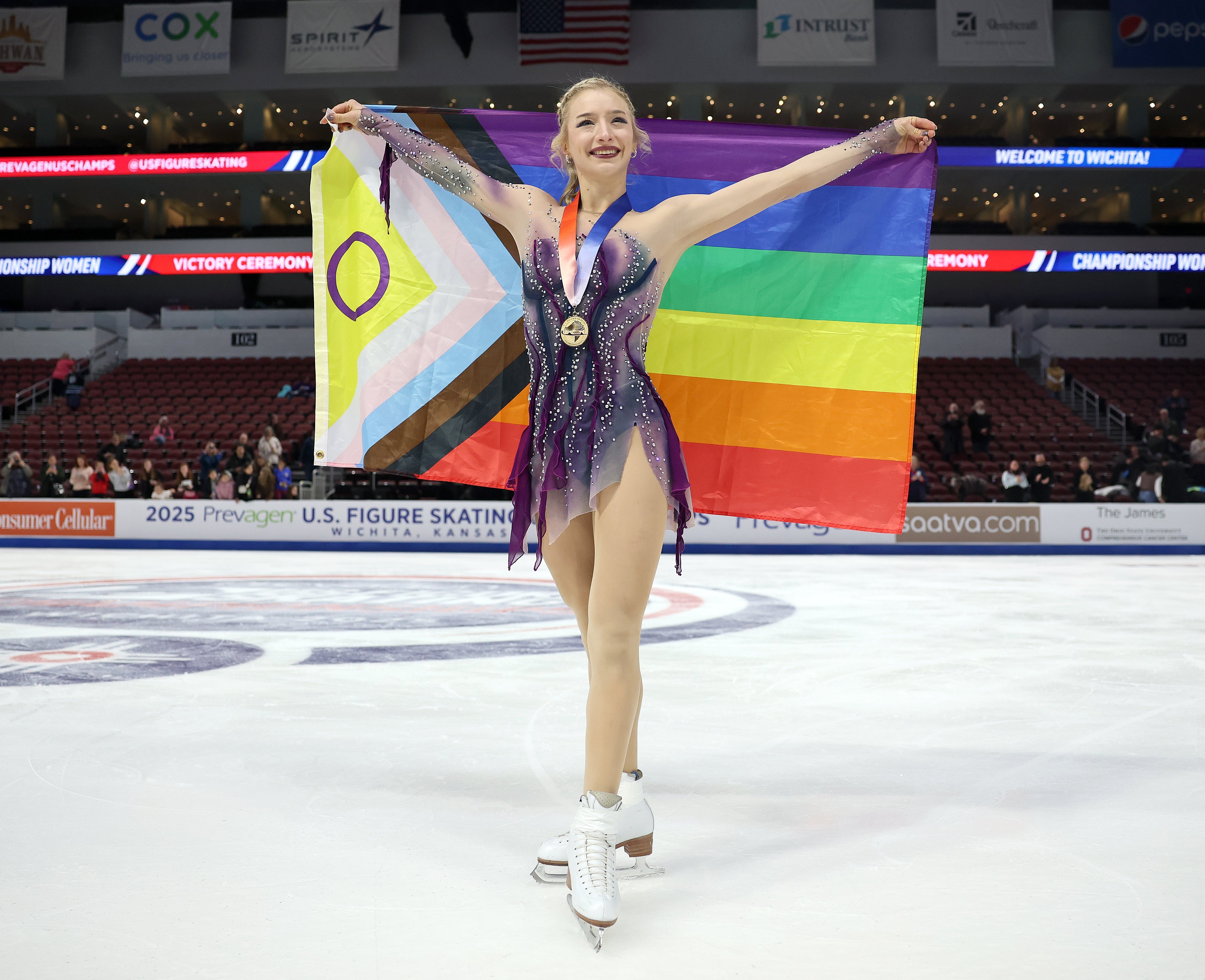 En kunstløper som har på seg et antrekk med paljetter holder et Pride-flagg og en medalje og smiler på en skøytebane under en konkurranseseremoni