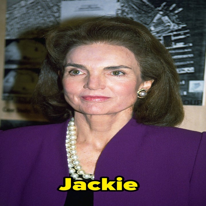 A person in a professional purple outfit with pearl necklace, standing against a background with black-and-white images