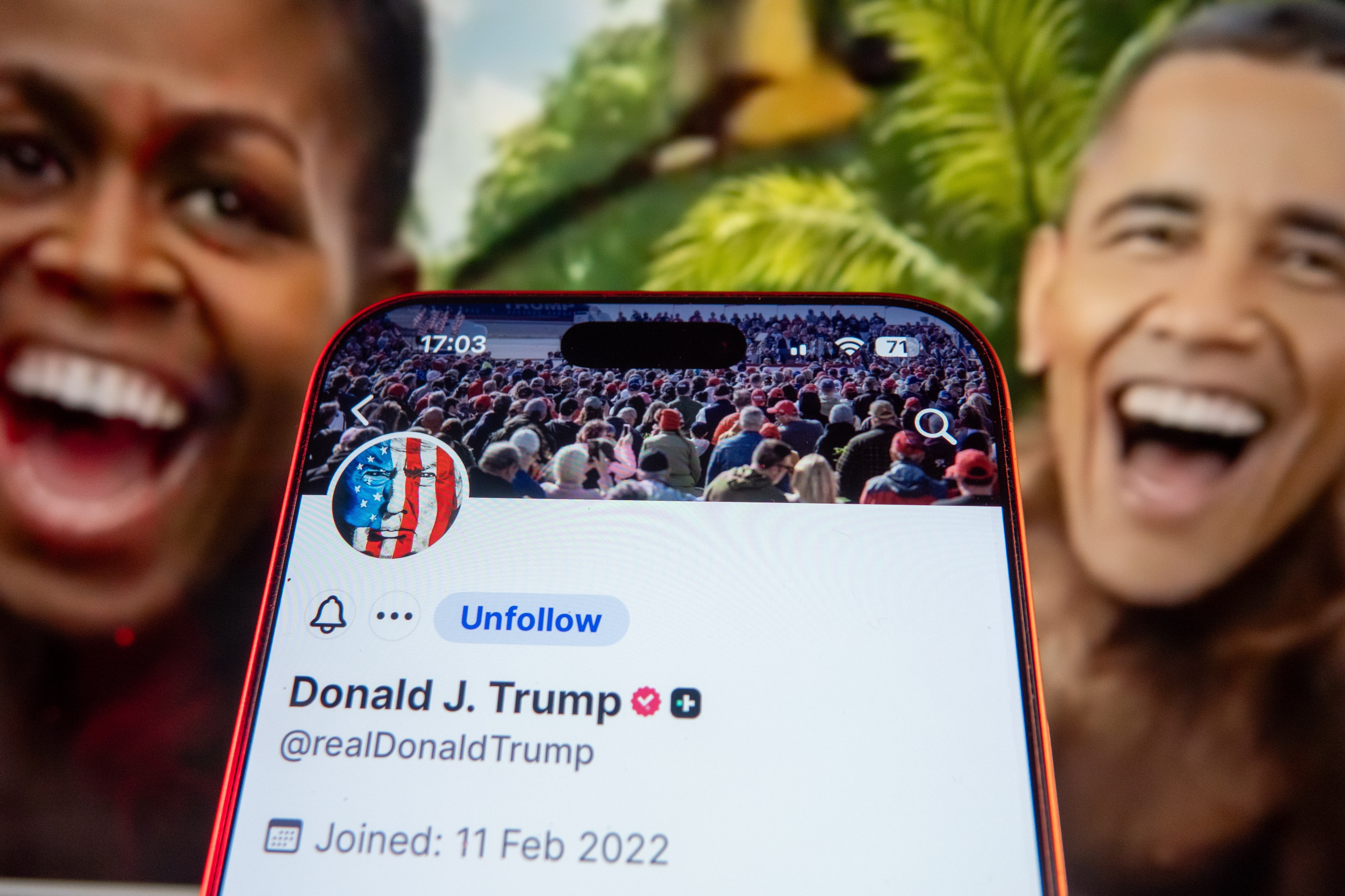 Smarttelefon viser Donald Trumps profil på sosiale medier foran uskarpt veggmaleri av smilende figurer med tropisk bakgrunn