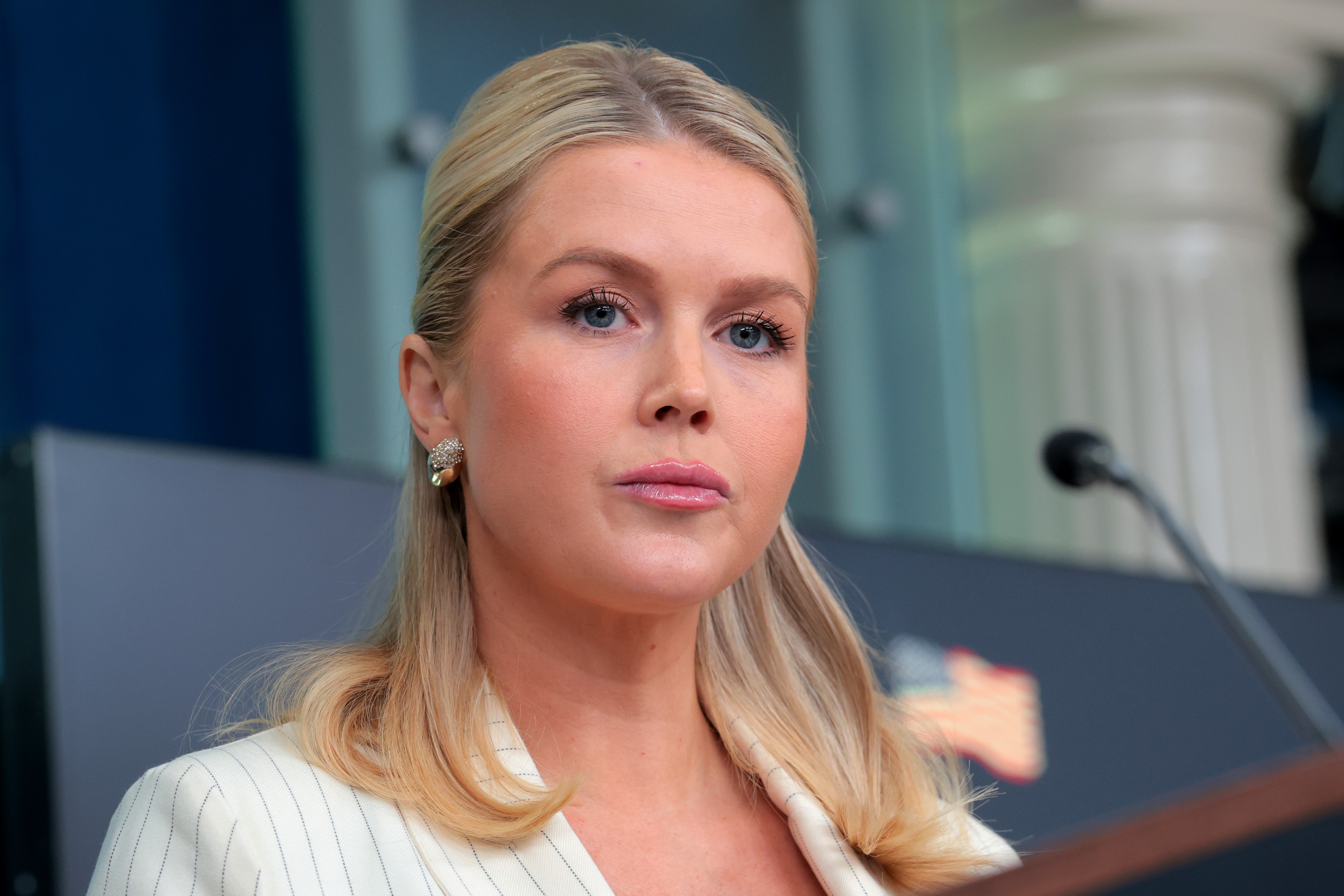 En person med langt hår snakker på et podium under en formell begivenhet, iført en pinstripe dress