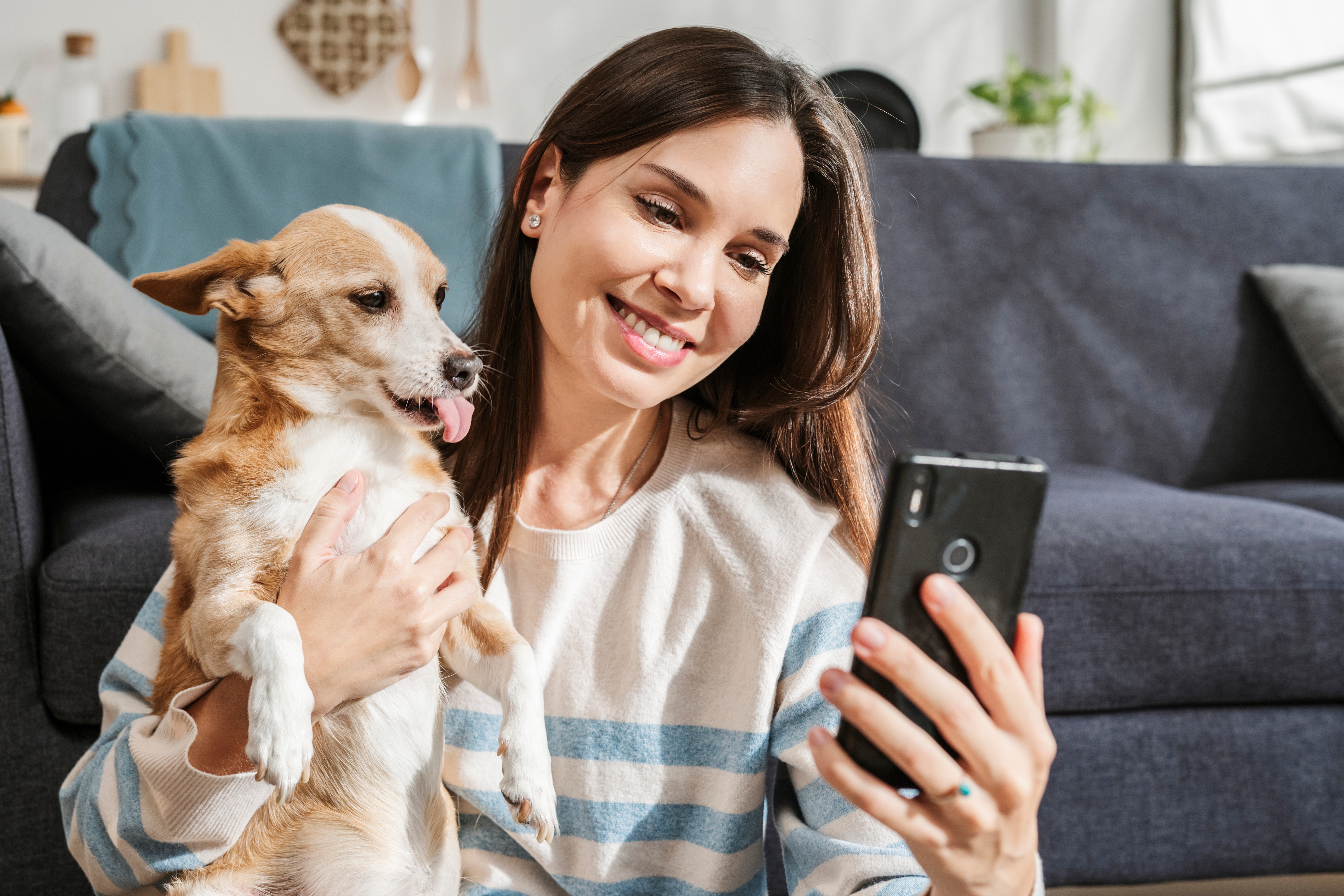 Woman smiling and holding her dog while taking a selfie on a smartphone, sitting in a cozy living room
