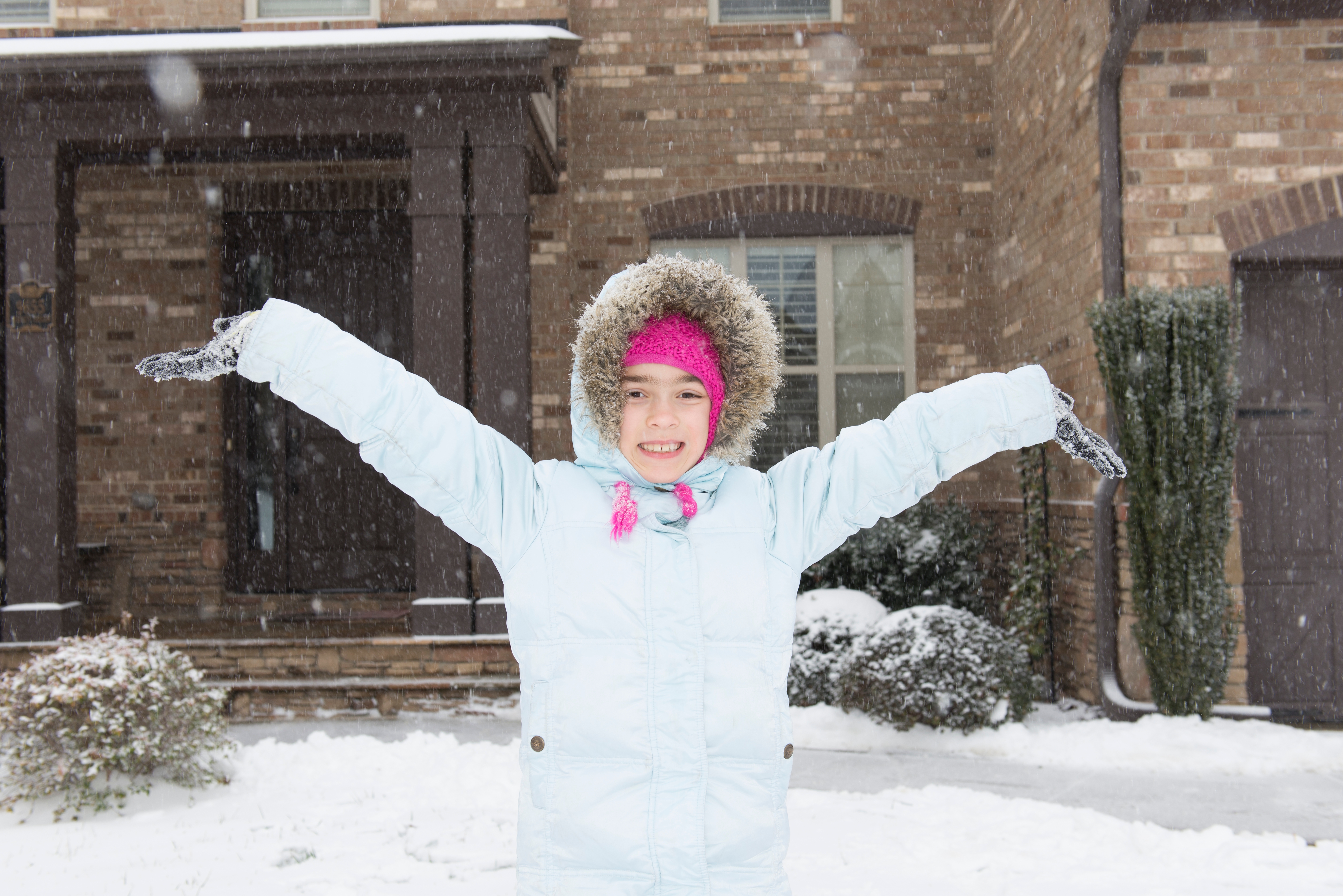 Child in winter coat and hat joyfully stands in falling snow, arms outstretched, in front of a brick house