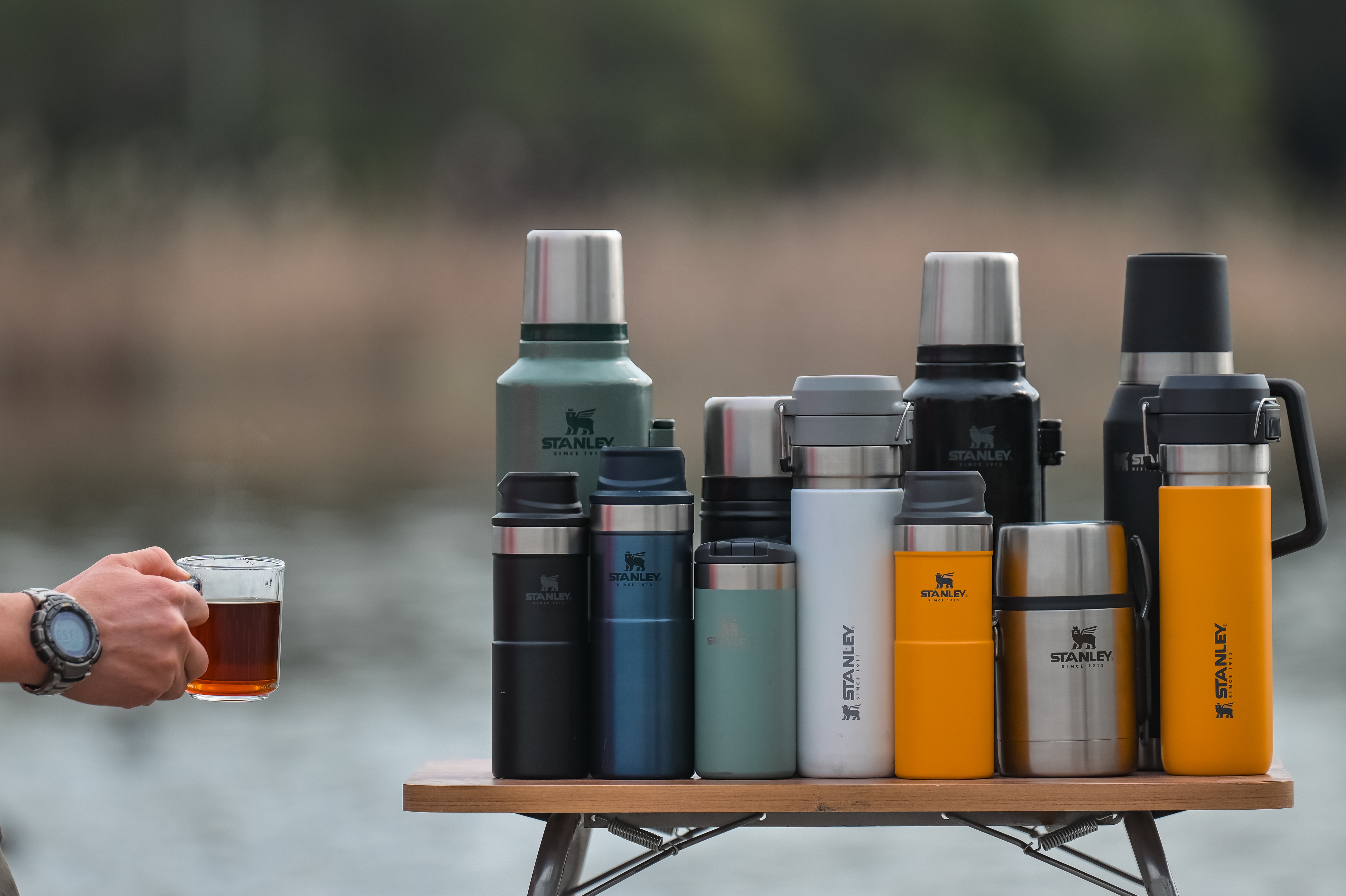 A person holding a metal mug filled with tea is standing next to a table with various Stanley thermoses of different sizes and styles