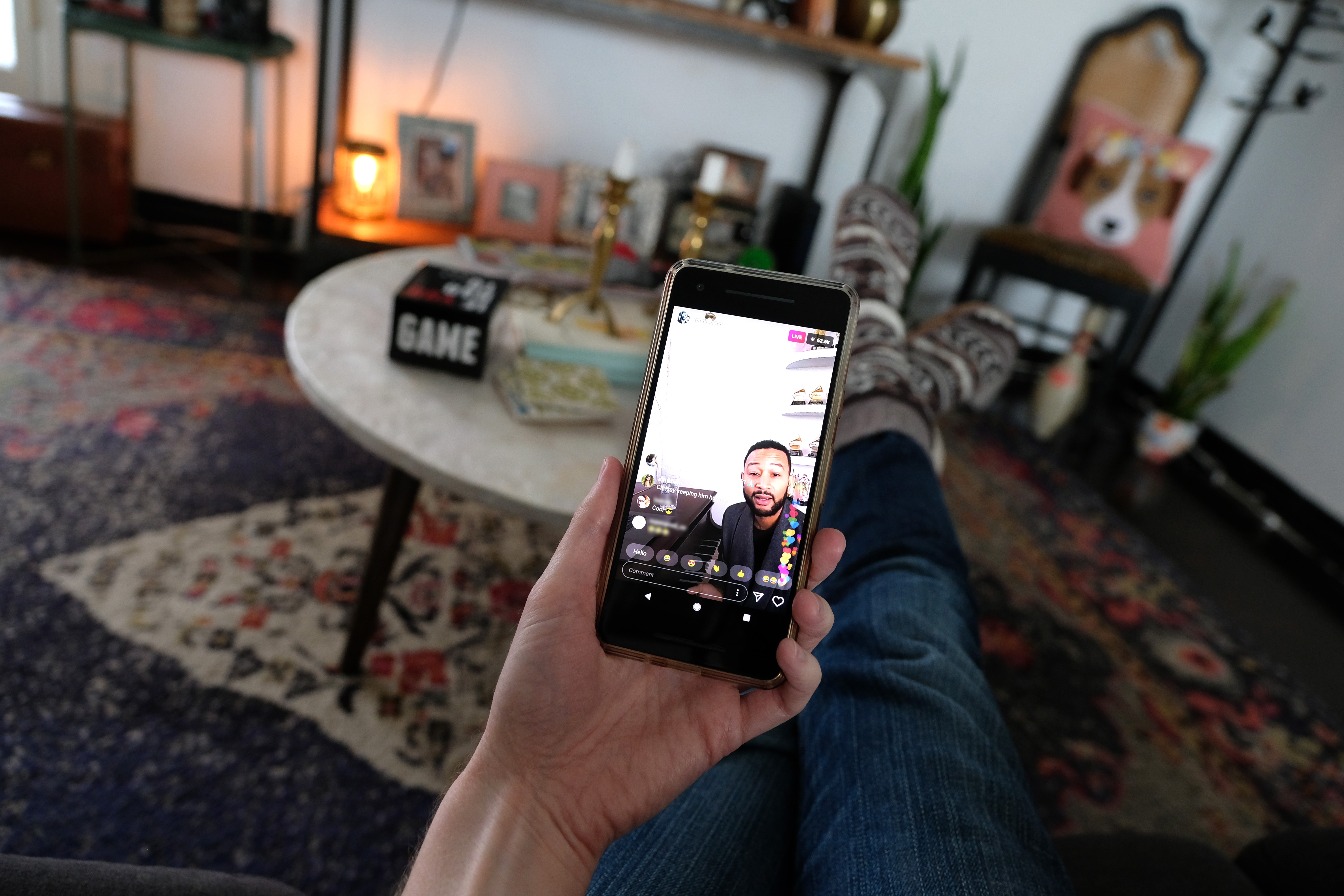 Person relaxing at home with feet up, watching a man speak in a livestream on a smartphone, surrounded by cozy room decor