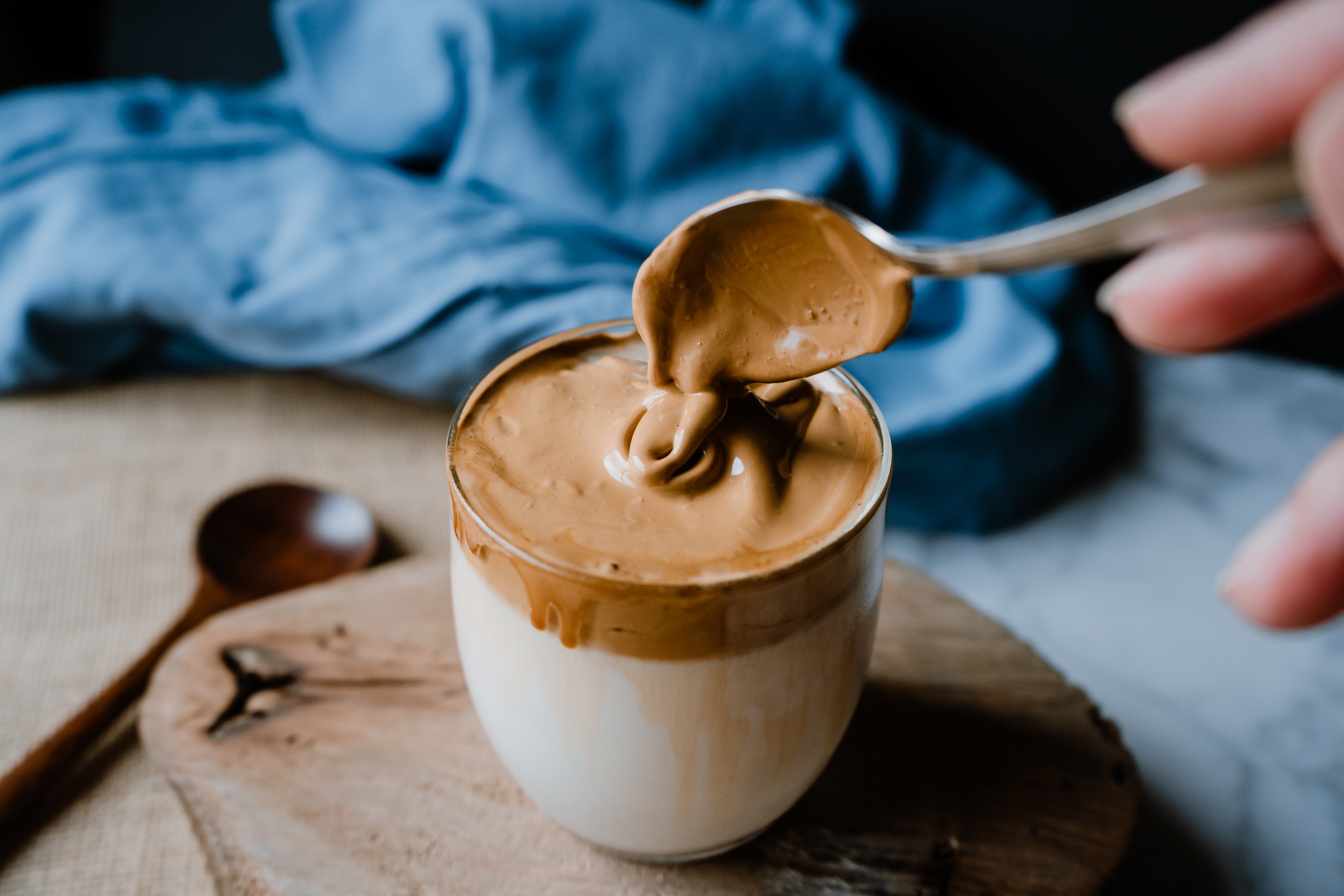 A hand holds a spoon with creamy dalgona coffee above a glass of milk on a wooden board, with a spoon in the background