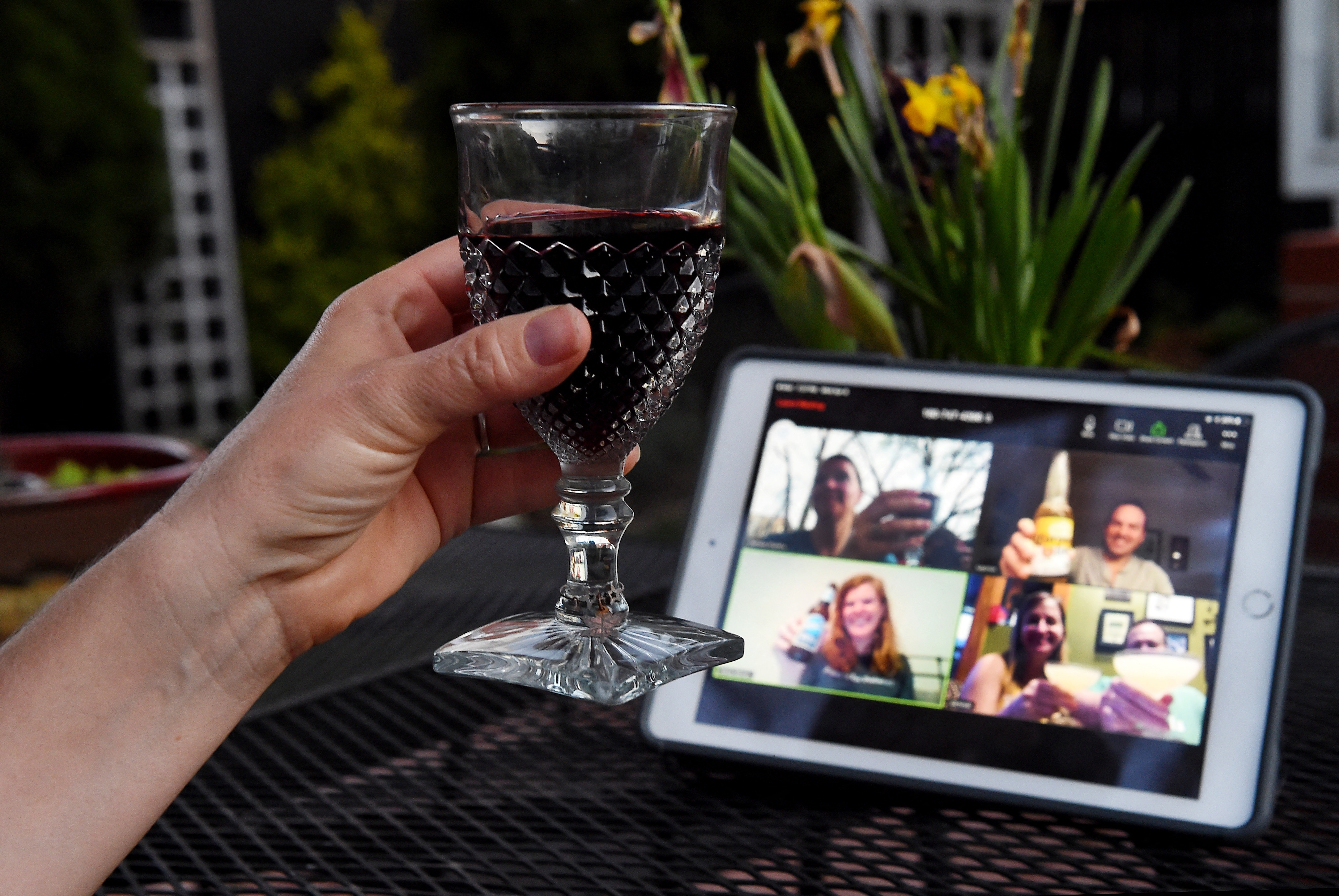 A person holds a wine glass during a video call on a tablet, with four people smiling on the screen, suggesting a virtual gathering