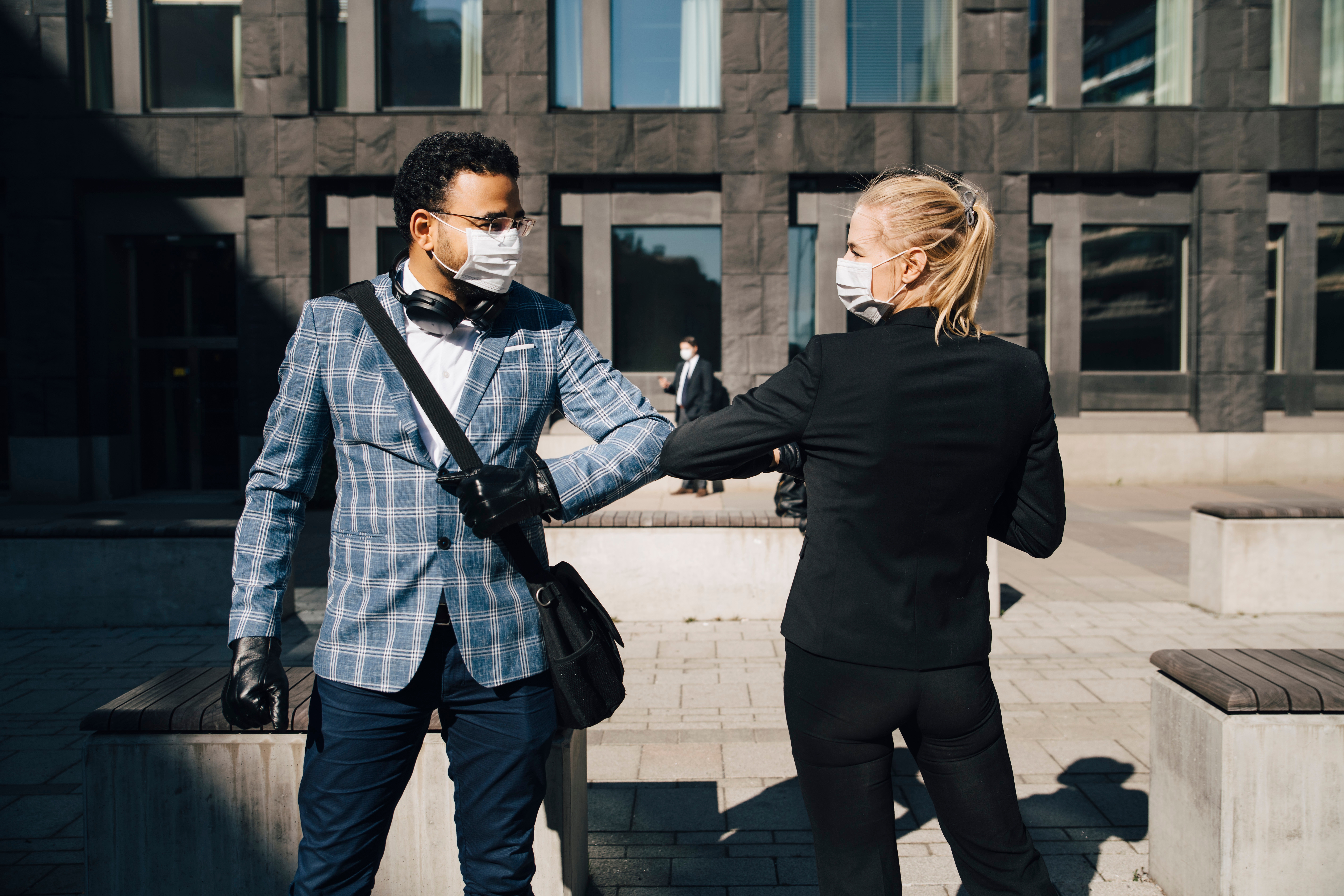 Two people in business attire wearing masks and gloves greet each other with an elbow bump outside a modern building