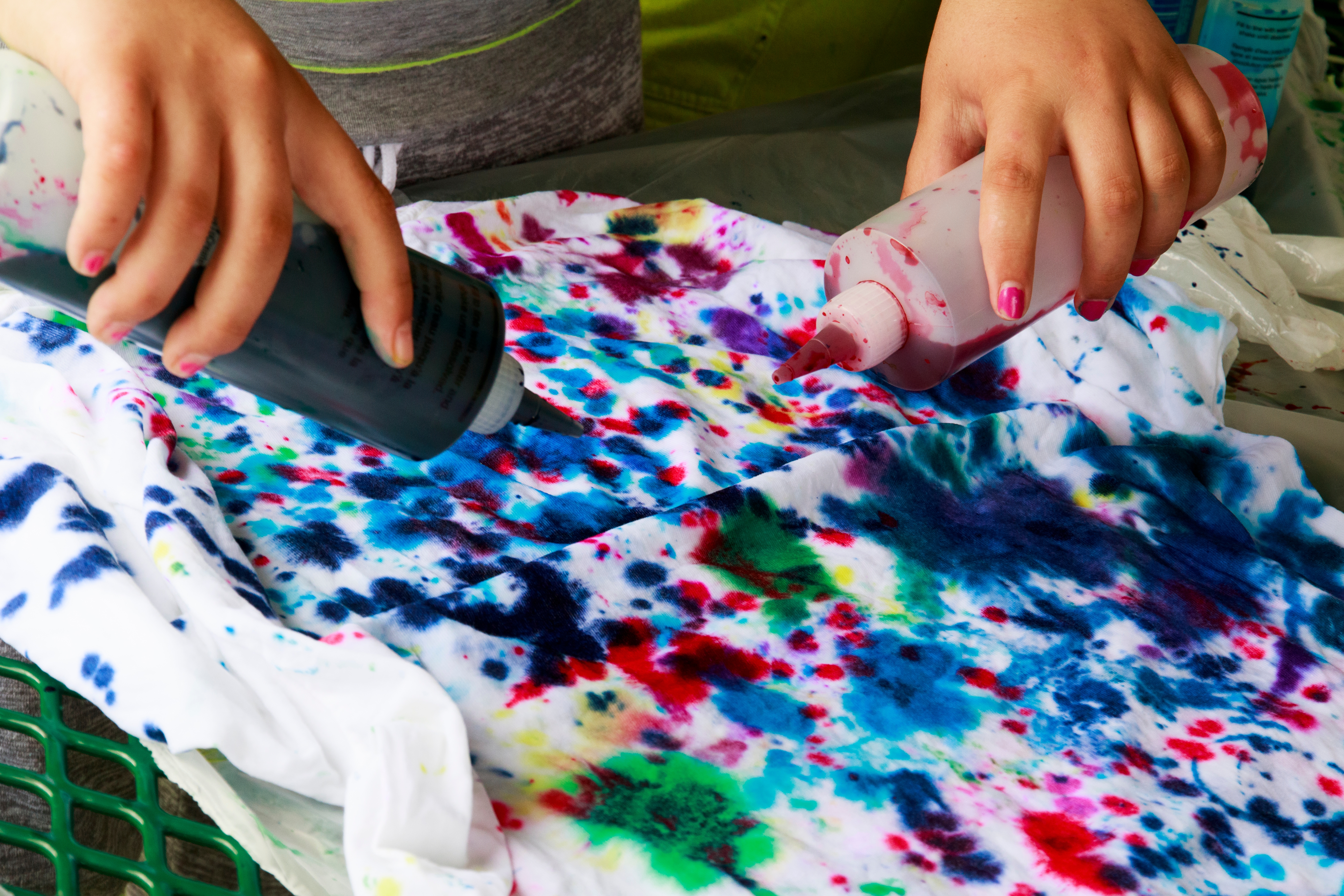 Person applying tie-dye to a shirt with squeeze bottles, creating a colorful pattern. Hands are visible, focusing on the creative process