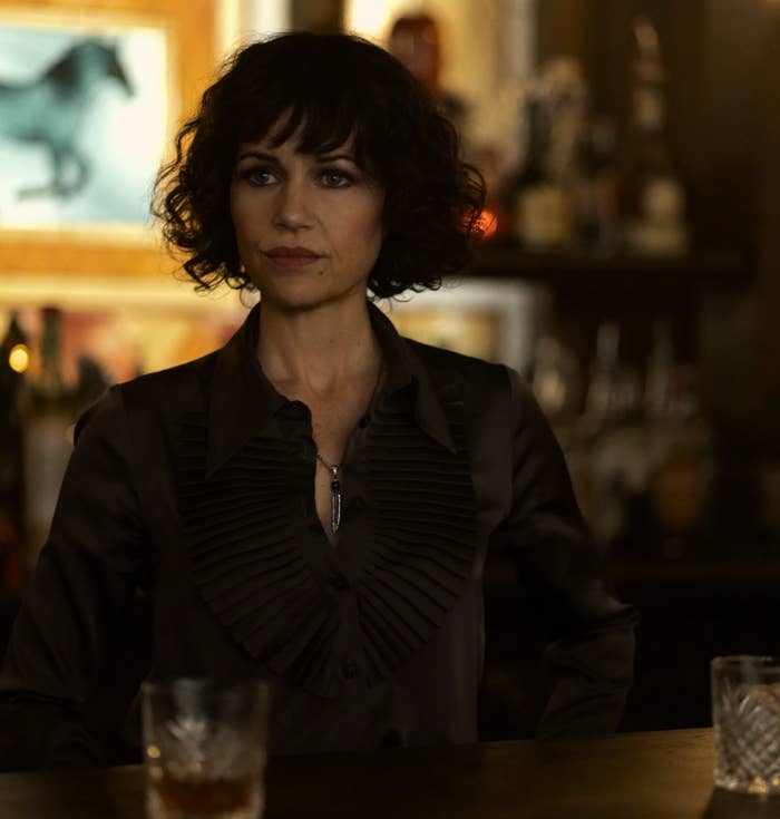 A idiosyncratic   with curly hairsbreadth  stands successful  a dimly lit bar, wearing a acheronian  garment  with ruffled detailing, looking thoughtful. A framed representation   is connected  the wall