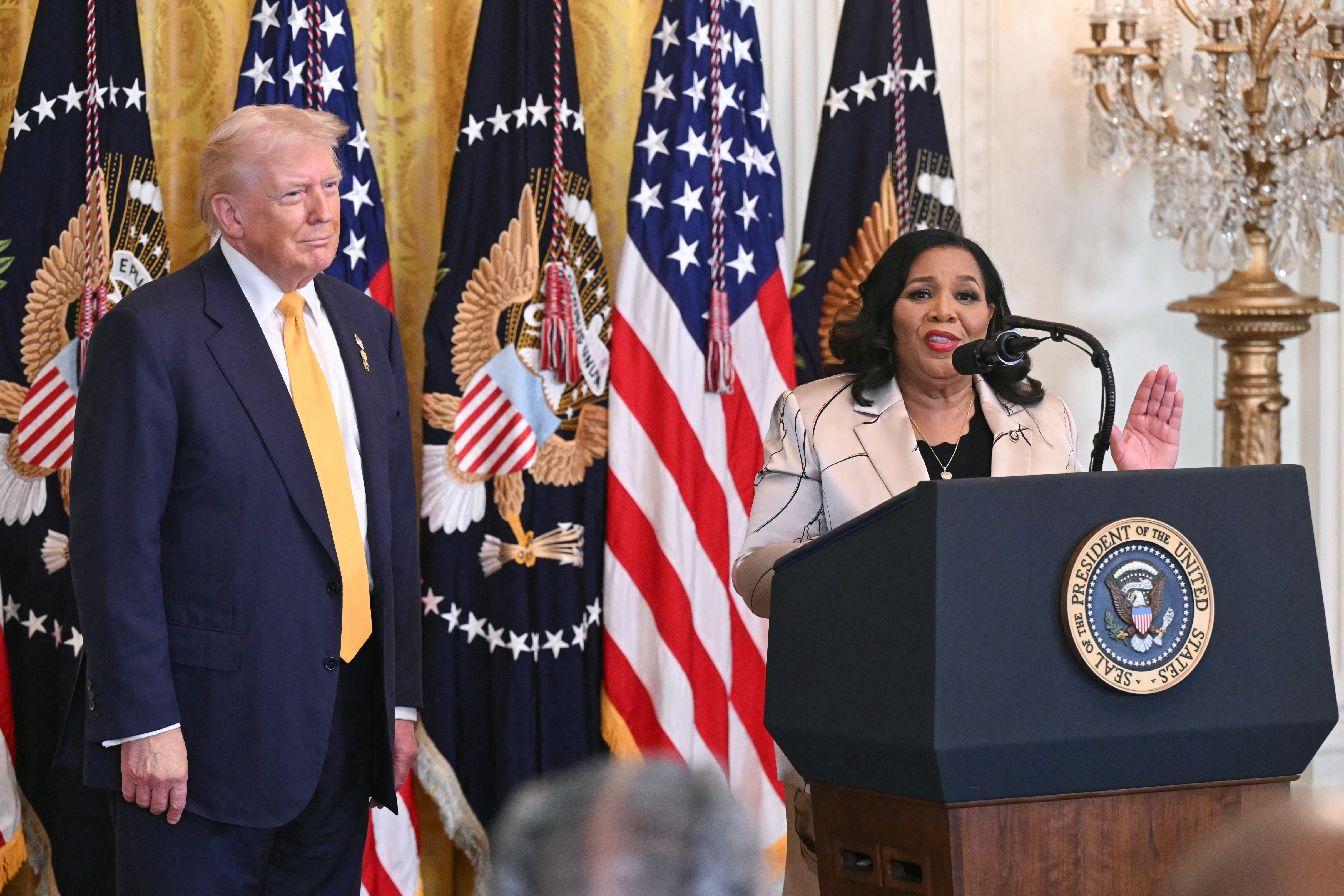 En person ved et podium snakker, med en passende mann som står i nærheten. De er i en formell setting med amerikanske flagg og presidentsegl