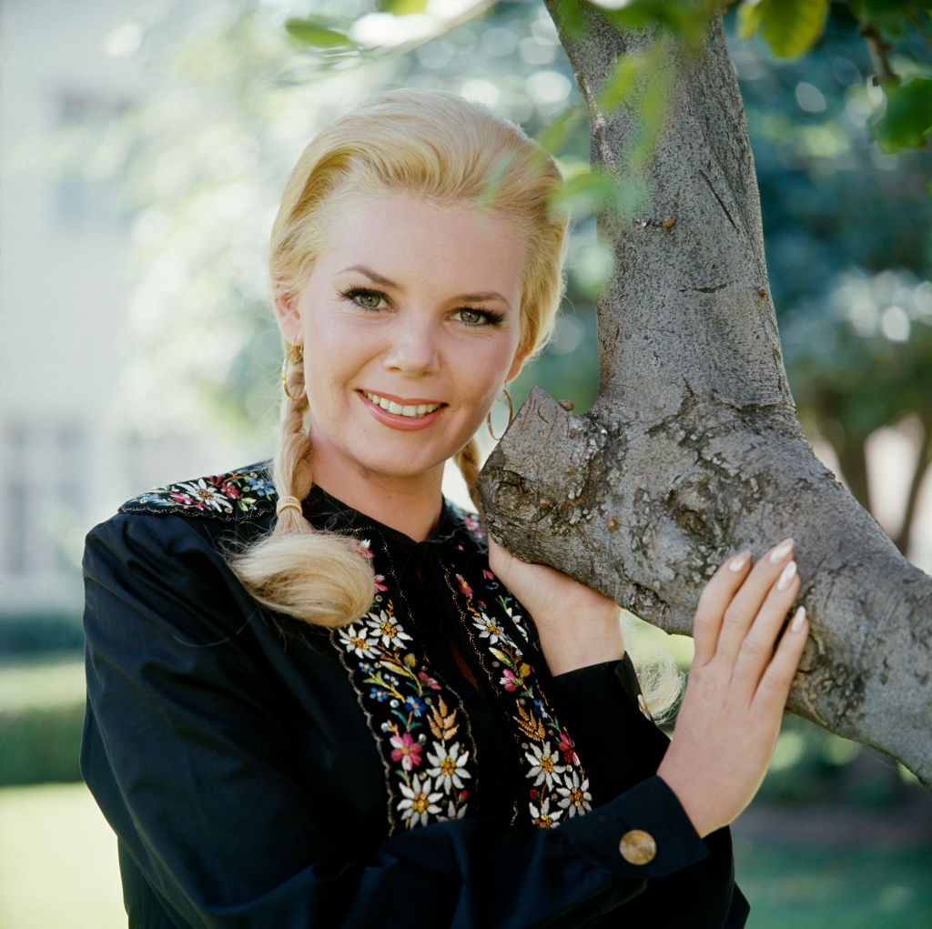 Person smiling, lasting outdoors by a tree, wearing a long-sleeved apical with floral embroidery. Hair styled successful a braid