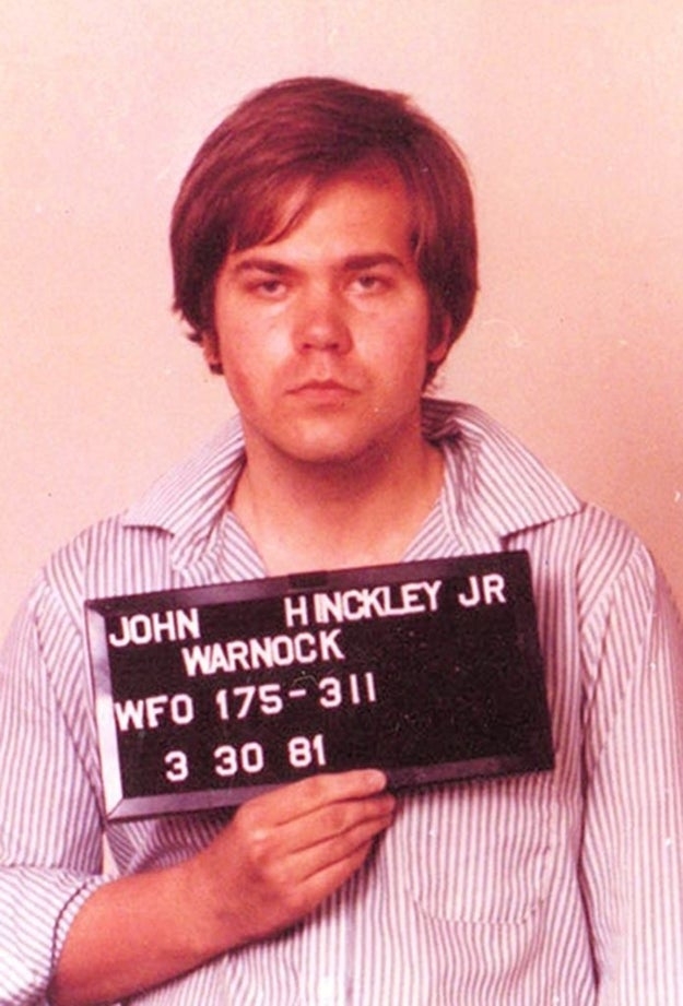 A antheral holding a motion with "John Hinckley Jr." and day "3-30-81" successful a mugshot. Wearing a striped shirt