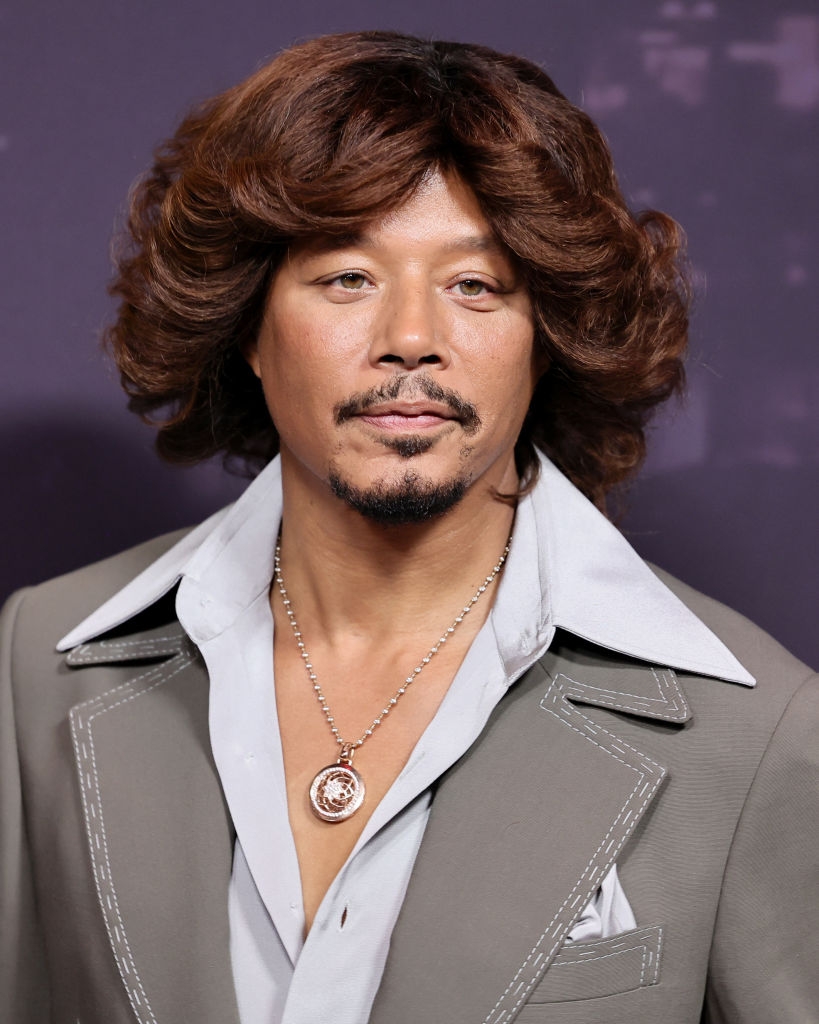 A idiosyncratic wearing a 70s-inspired blazer and garment with wide lapels, accessorized with a necklace, is posing astatine a personage event