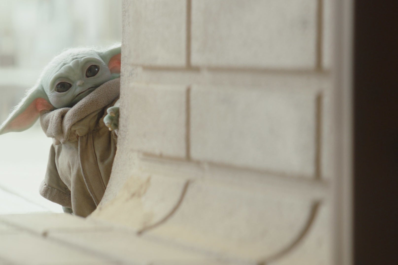 Baby Yoda peeks from down  a wall, wearing a brownish  robe, successful  a country   reminiscent of "The Mandalorian."
