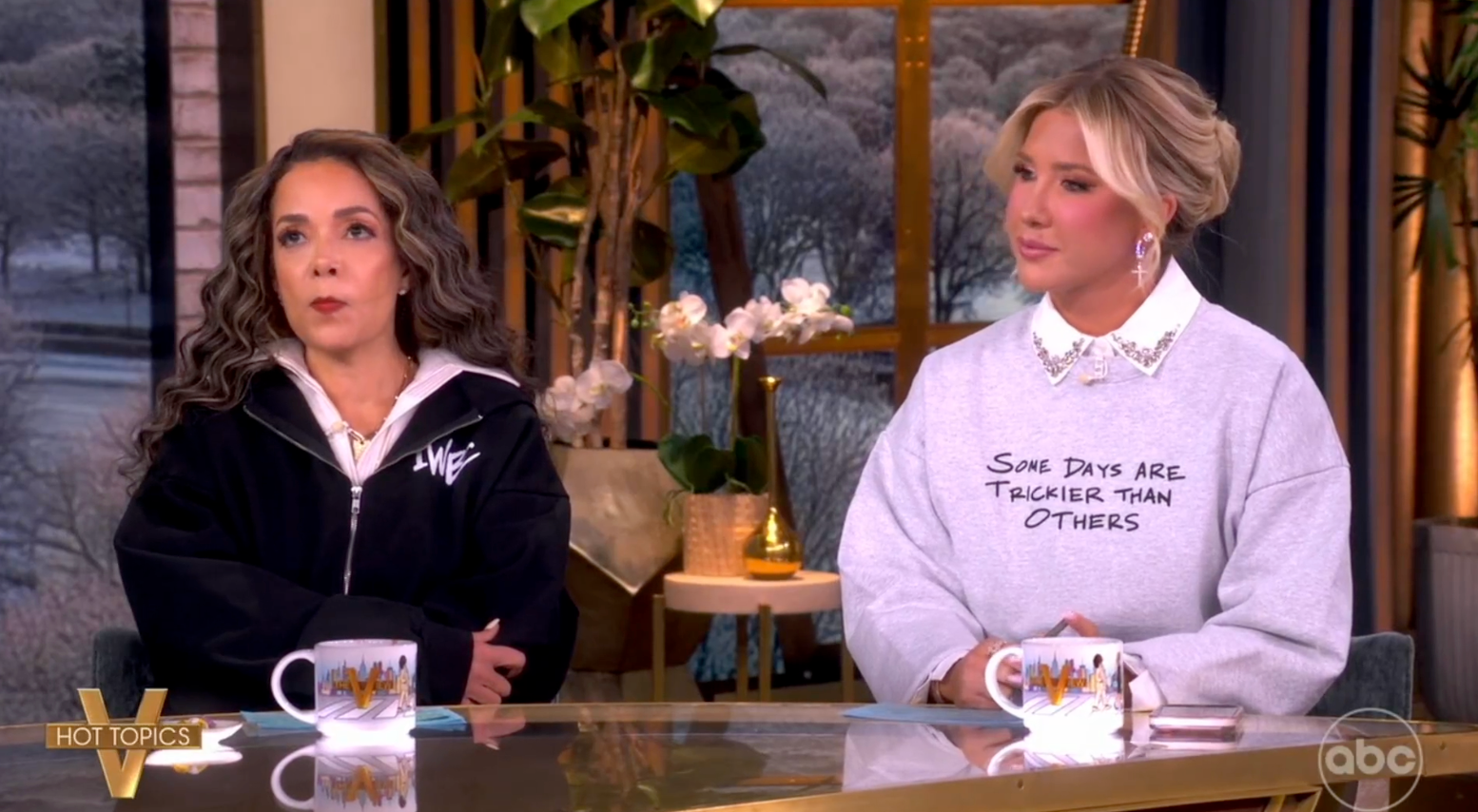 Two women beryllium   astatine  a speech   amusement   table. One wears a zipped jacket; the different   wears a sweatshirt with text. They person  mugs successful  beforehand   of them