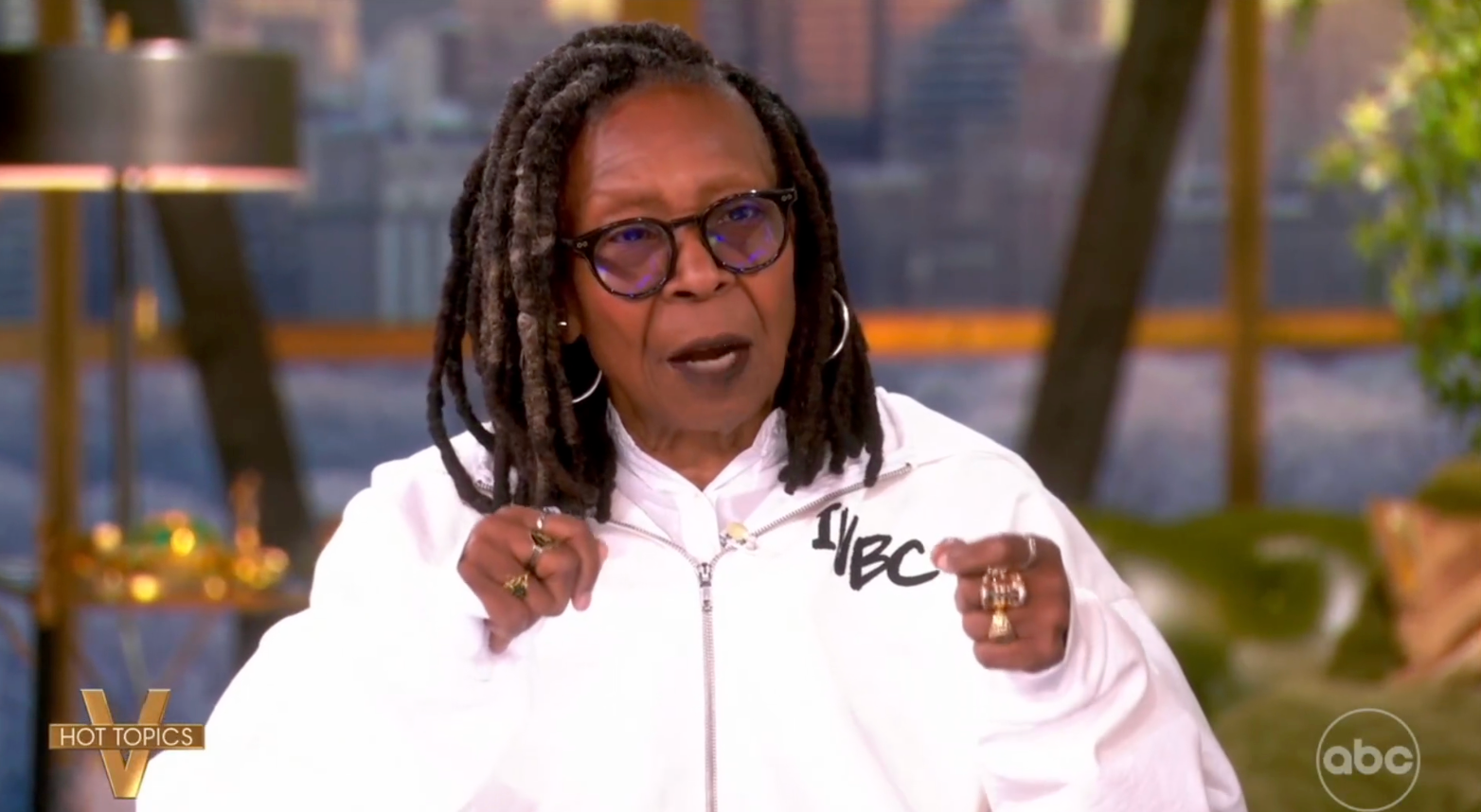 Person med dreadlocks iført briller og en hvit jakke med tekst, snakker på et sett med et bybilde i bakgrunnen