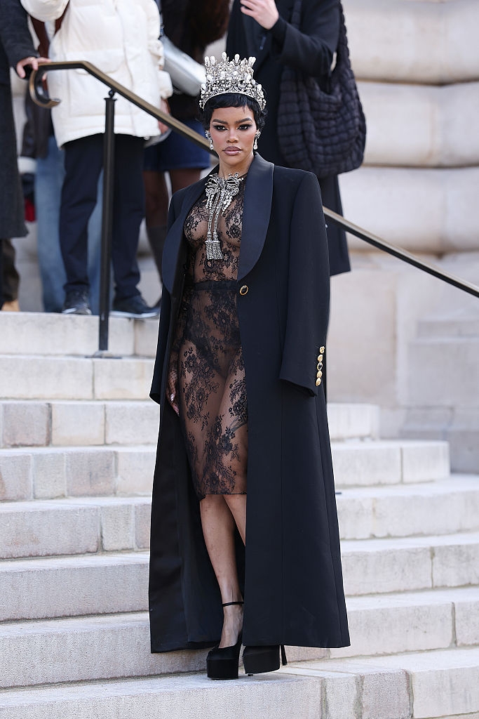 Person successful a agelong achromatic overgarment implicit a lace dress, adorned with an ornate crown and necklace, stands confidently connected steps successful a ceremonial setting
