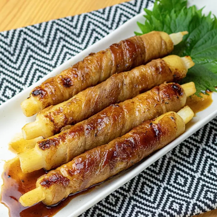 白いアスパラガスに肉が巻かれ、照り焼きソースがかかった料理。葉が添えられている。