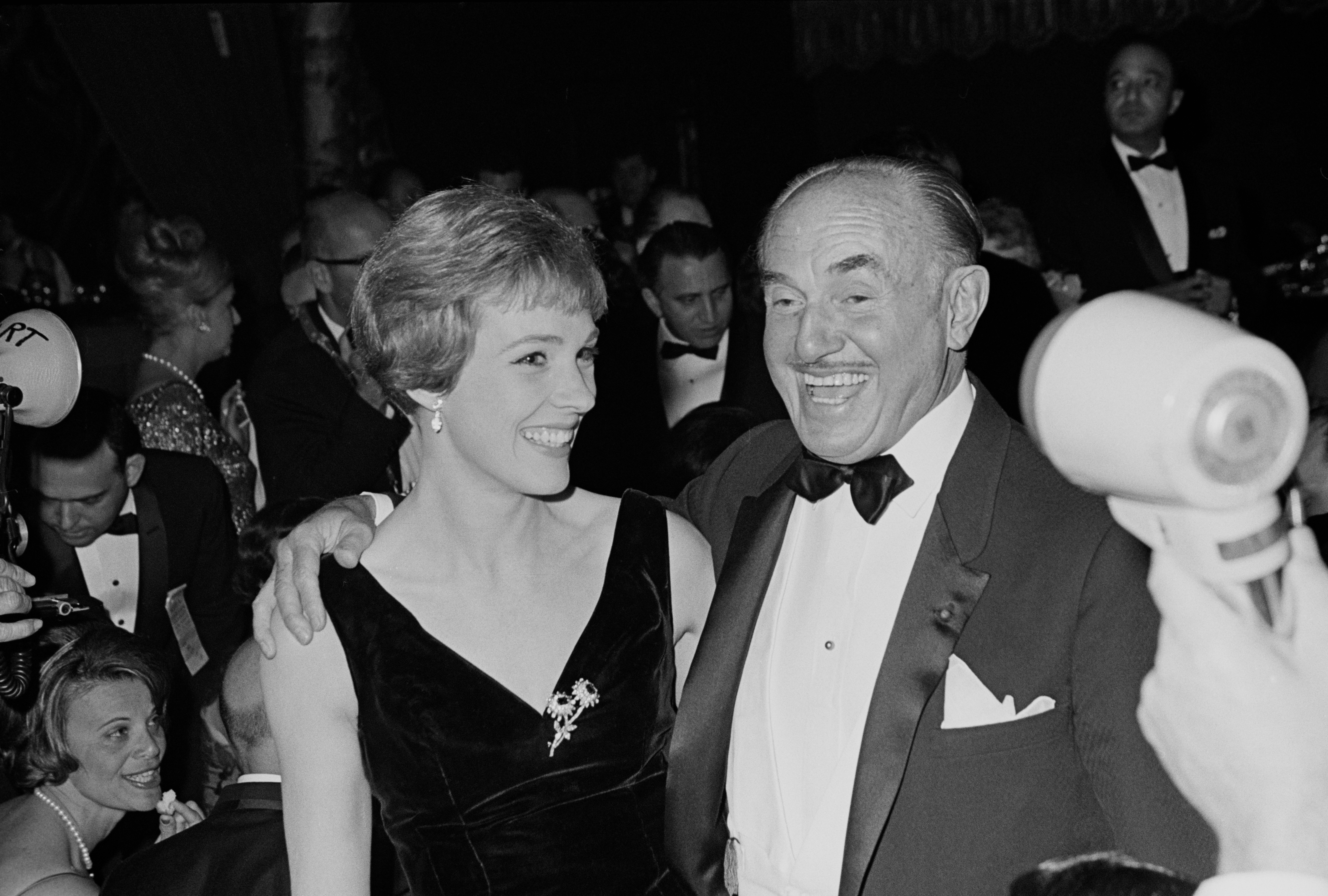 A pistillate   successful  an elegant sleeveless formal  smiles beside a antheral   successful  a tuxedo astatine  a ceremonial  event. The inheritance  shows different   guests successful  evening attire