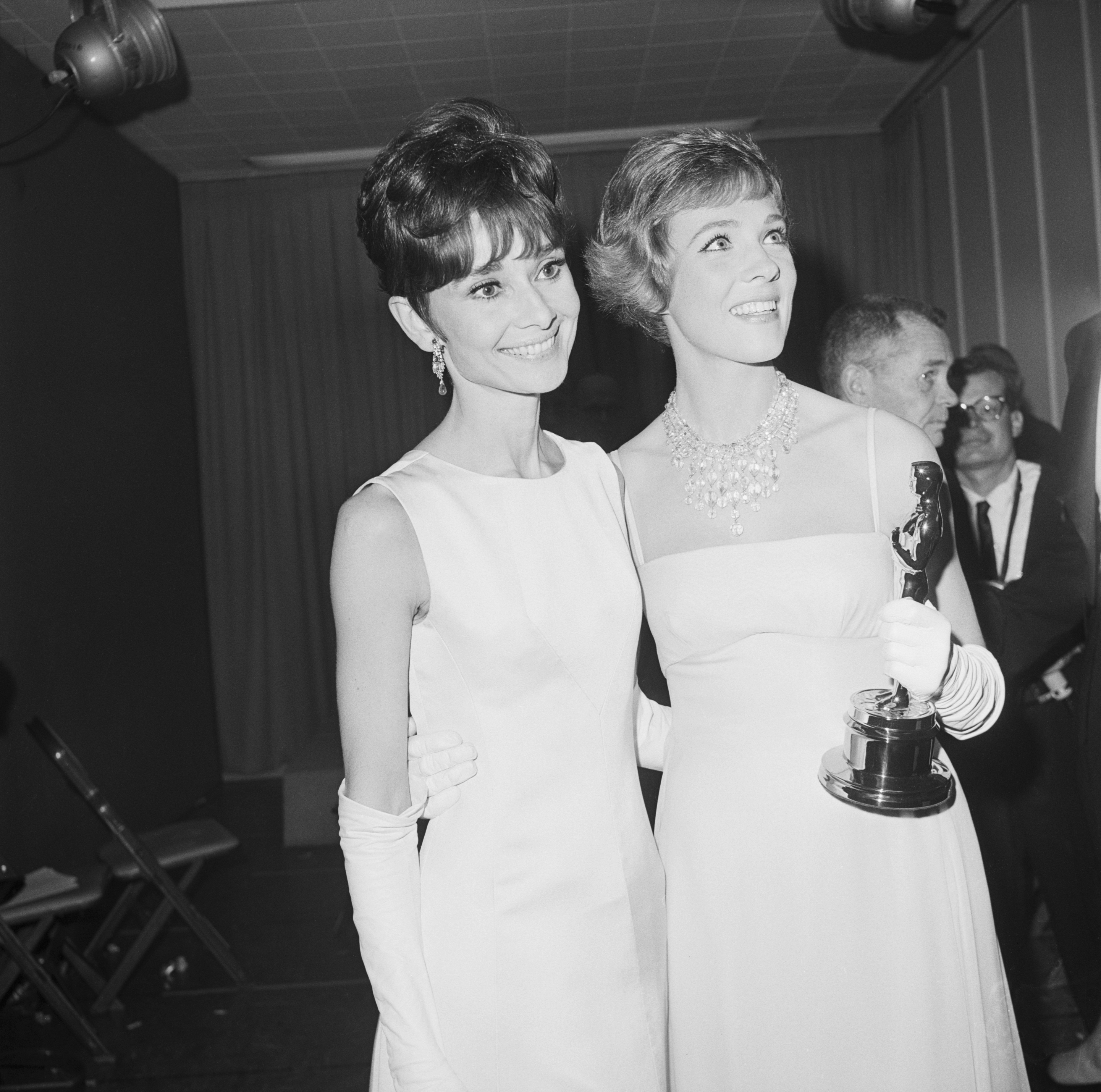 Two women successful  elegant gowns holding an award, smiling joyfully. One pistillate   wears gloves and a connection    necklace