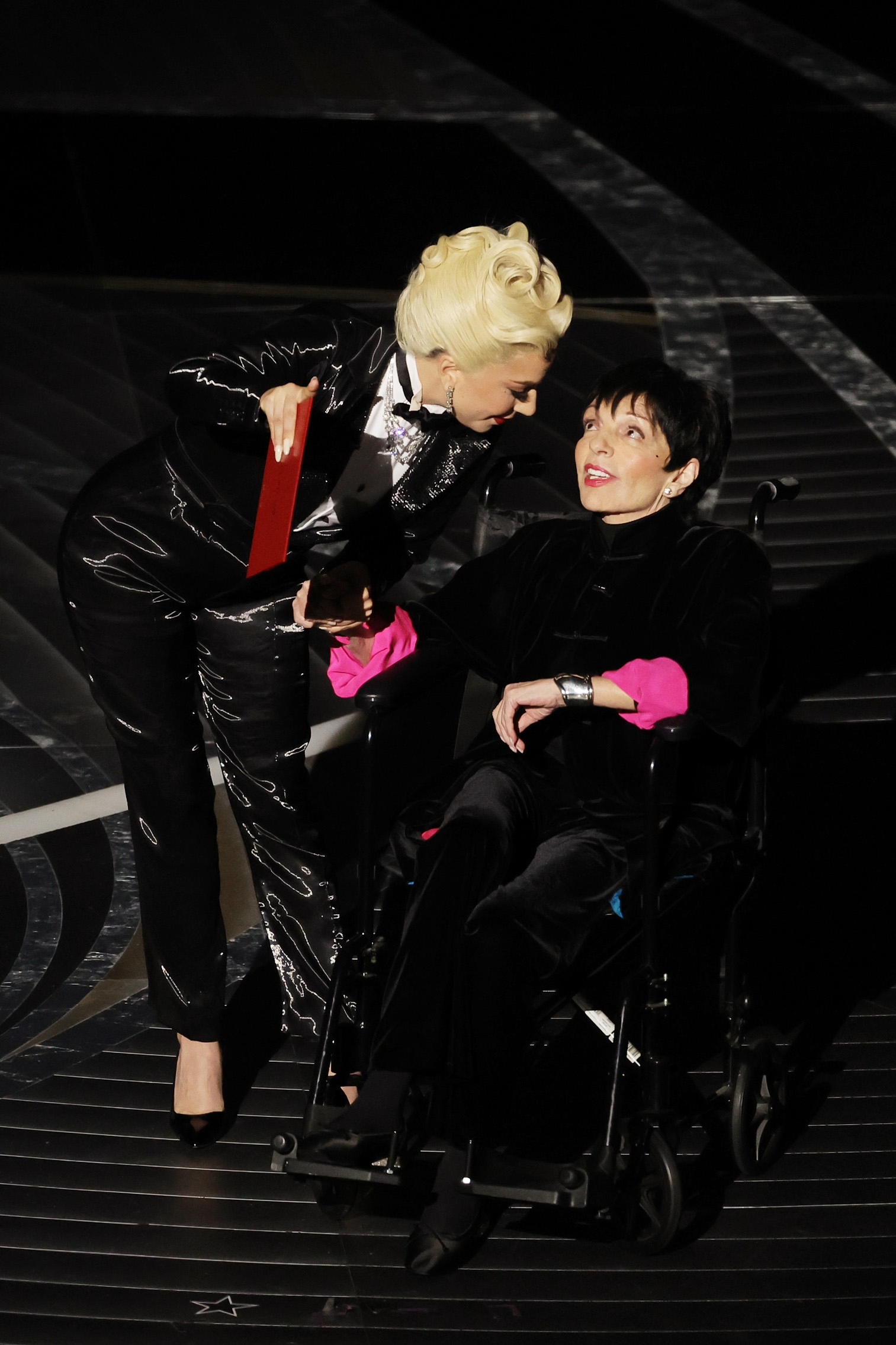 Lady Gaga lasting  successful  a shiny tuxedo suit   and Liza Minnelli seated successful  a wheelchair, sharing a moment