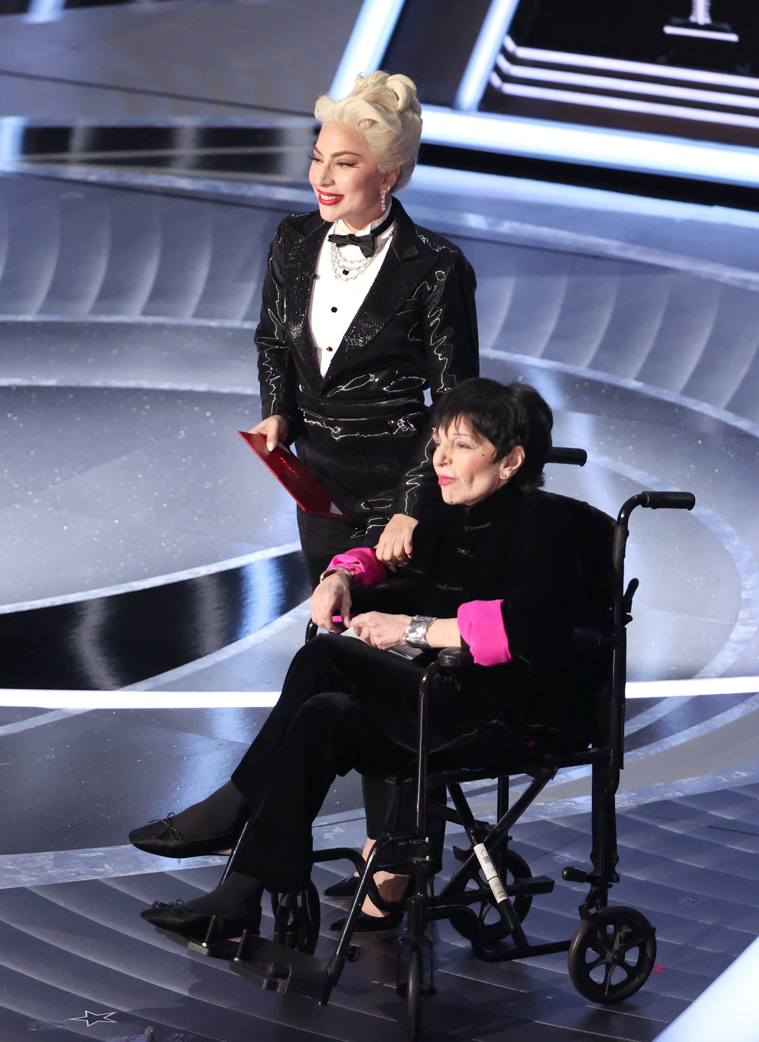 Lady Gaga successful  a tuxedo suit   holding an envelope and Liza Minnelli sitting successful  a wheelchair wearing an outfit wiith heavy   cuffs