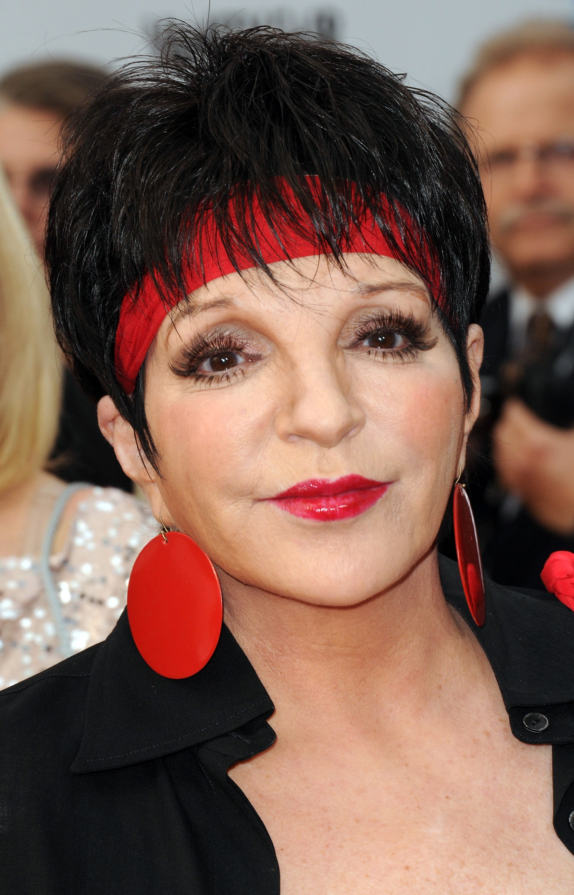 Liza Minnelli with abbreviated  hair, striking lipstick, and ample  reddish  earrings, wearing a headband and blouse connected  a reddish  carpet