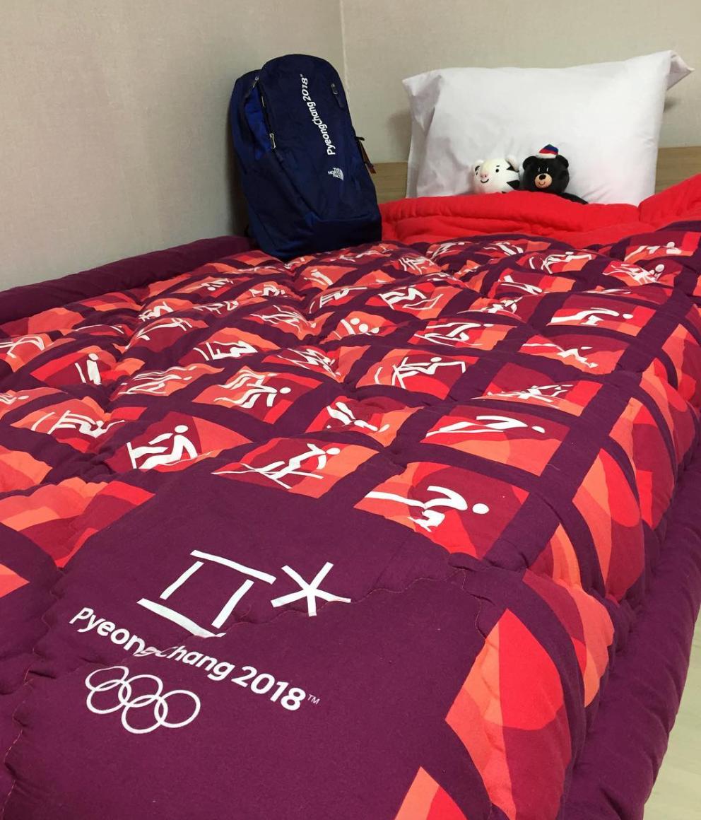 En seng har et PyeongChang 2018 olympisk teppe med sportsikoner. En blå ryggsekk og to plysjleker ligger på puten