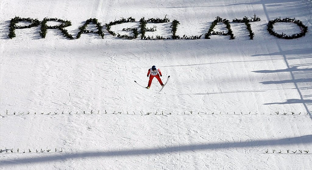 Skiløper konkurrerer på en snødekt bane under store "PRAGELATO" bokstaver laget av grønt, kaster skygger på snøen