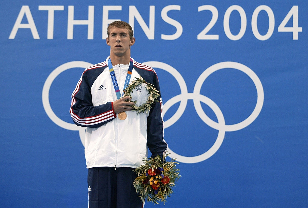Idrettsutøver på pallen ved OL i Athen 2004, iført treningsdress og holder blomster og medalje, med olympiske ringer i bakgrunnen