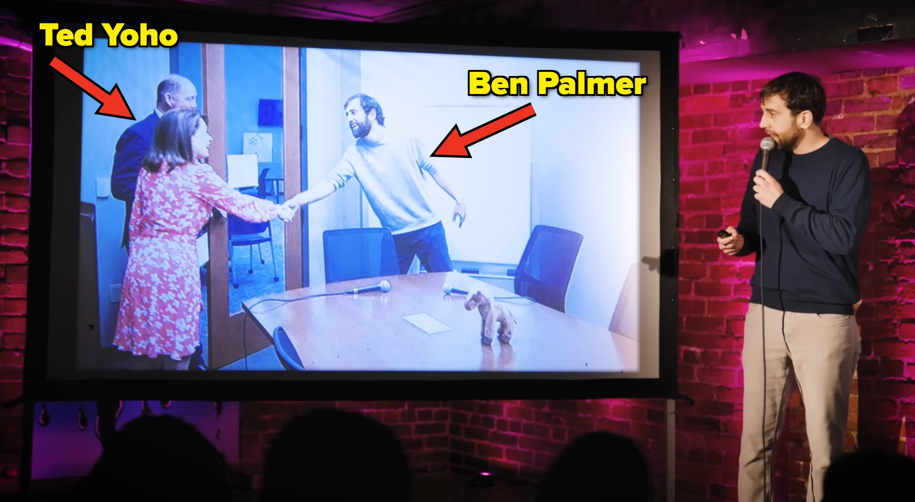 En mann med en mikrofon presenterer et projisert bilde av tre personer i et kontormiljø, engasjert i et håndtrykk