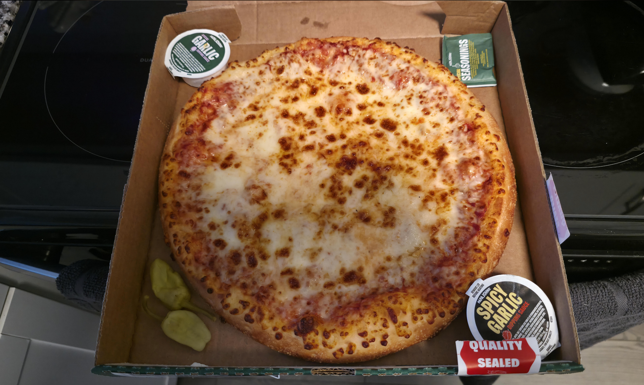 A freshly baked cheese pizza in a delivery box with garlic sauce cups and pepperoncini on the side
