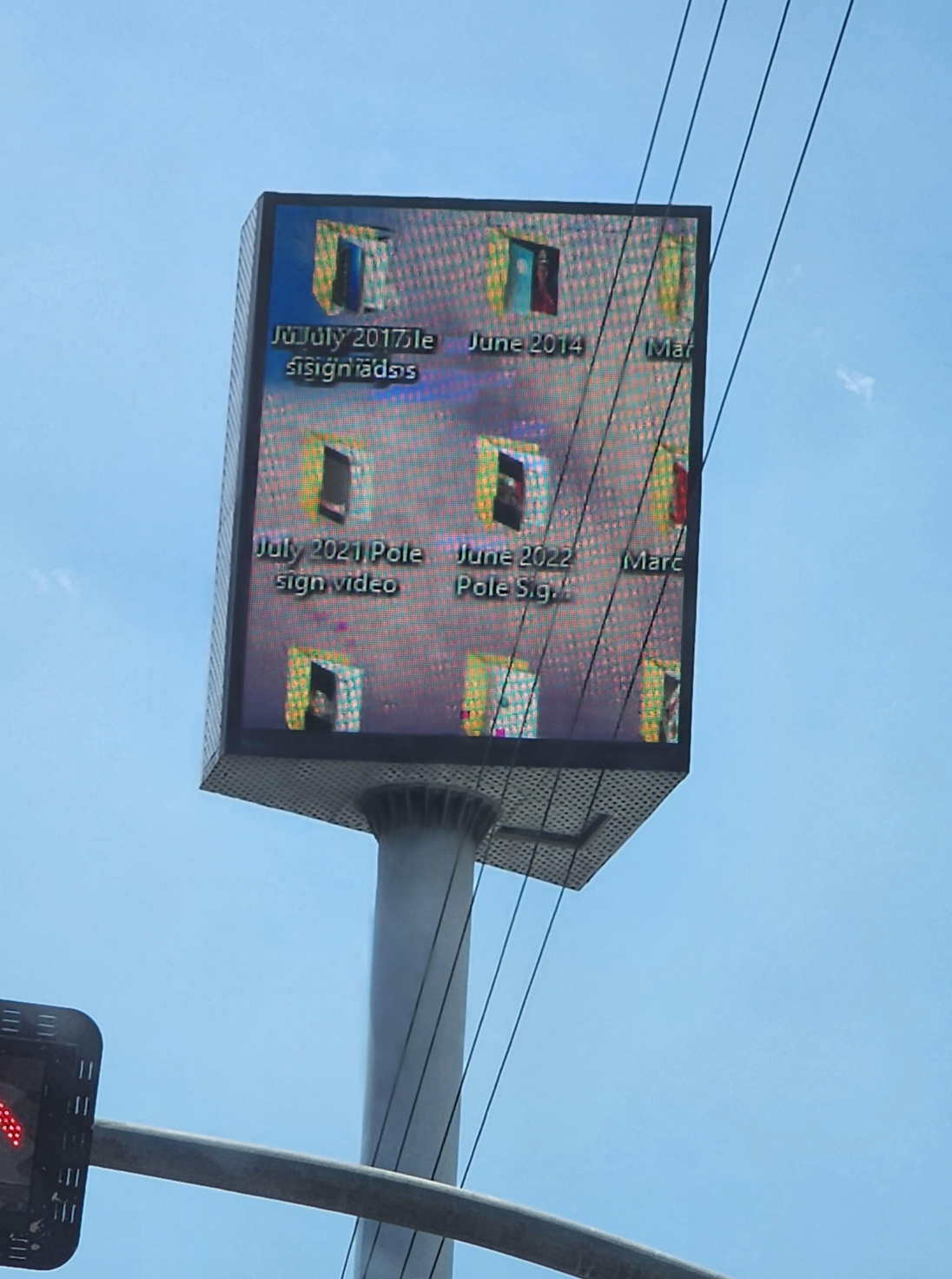 Traffic light with a pixelated electronic sign displaying file names like "July 2021 Pole Sign Video" against a clear sky