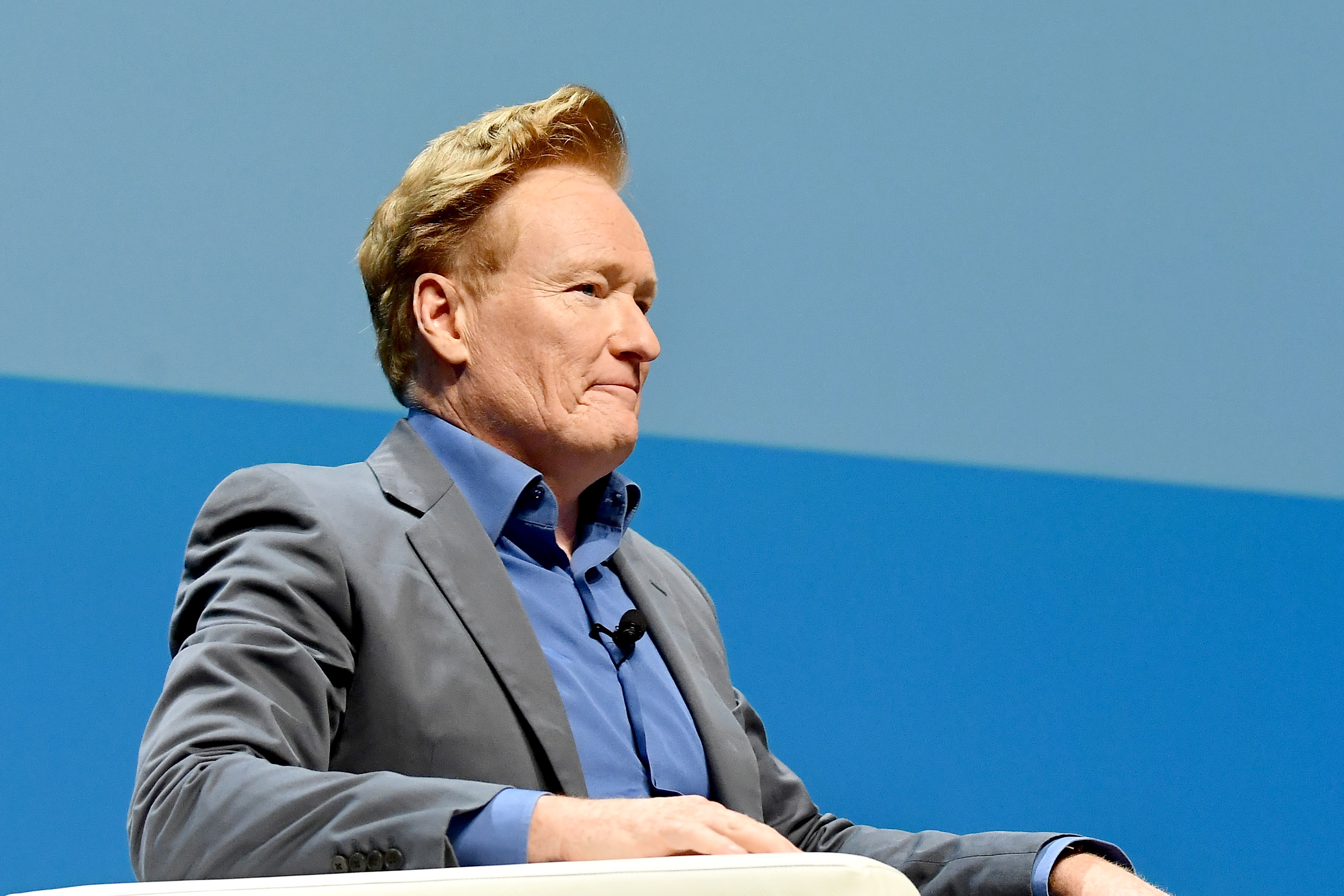 Conan O'Brien i dress sitter på scenen, virker engasjert i tanker eller lytter oppmerksomt