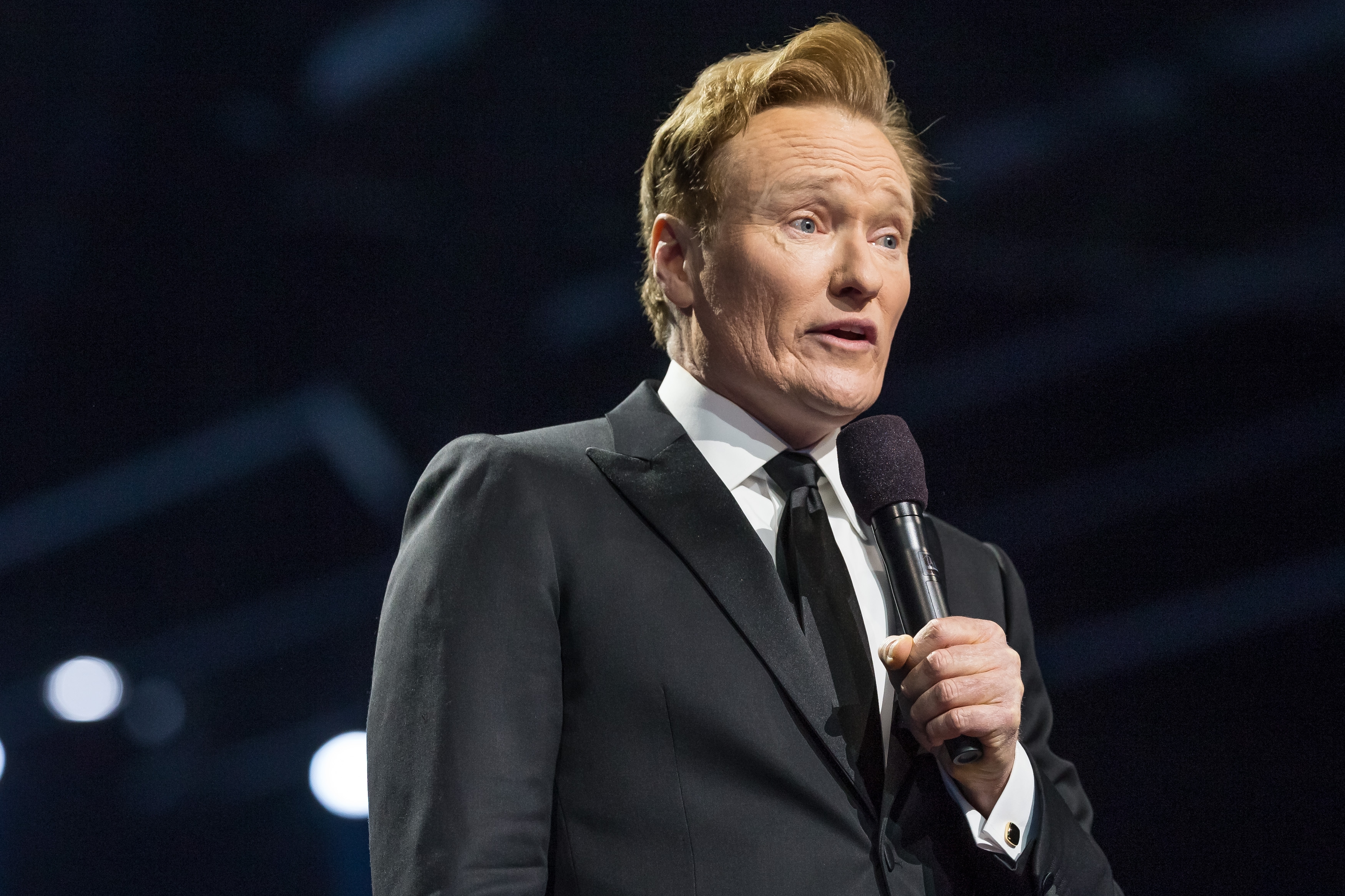 Conan O'Brien i dress holder en mikrofon, snakker på scenen ved et offentlig arrangement
