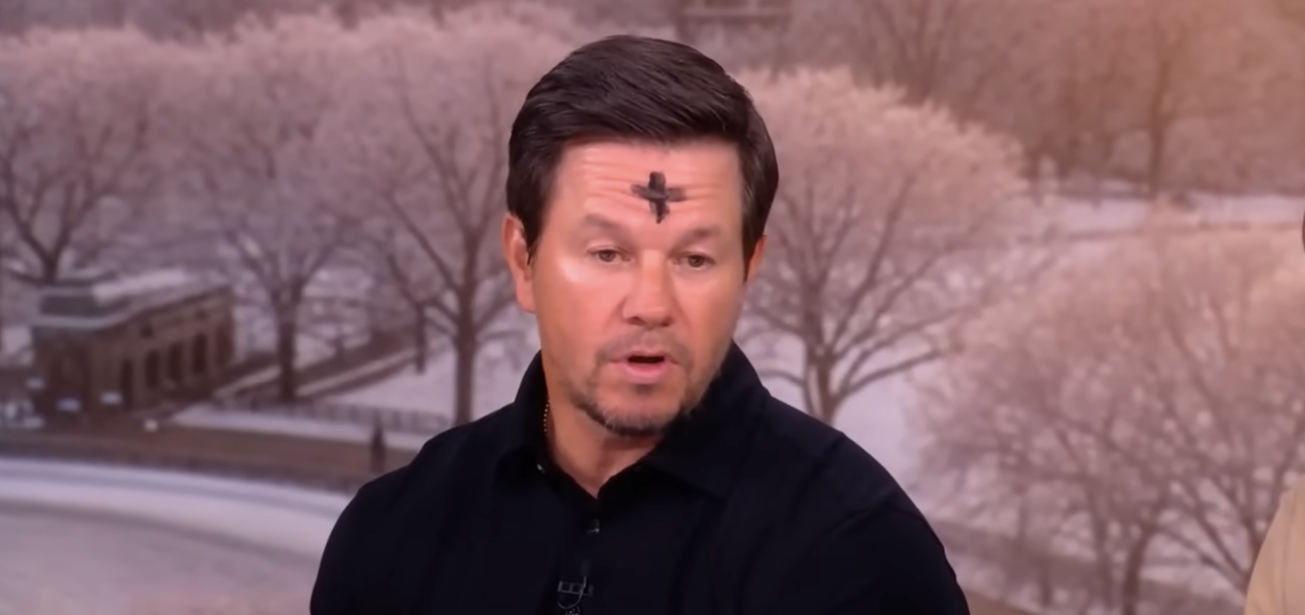 Man speaking on TV show with an ash cross on his forehead, wearing a black shirt. Snowy park scene visible in background