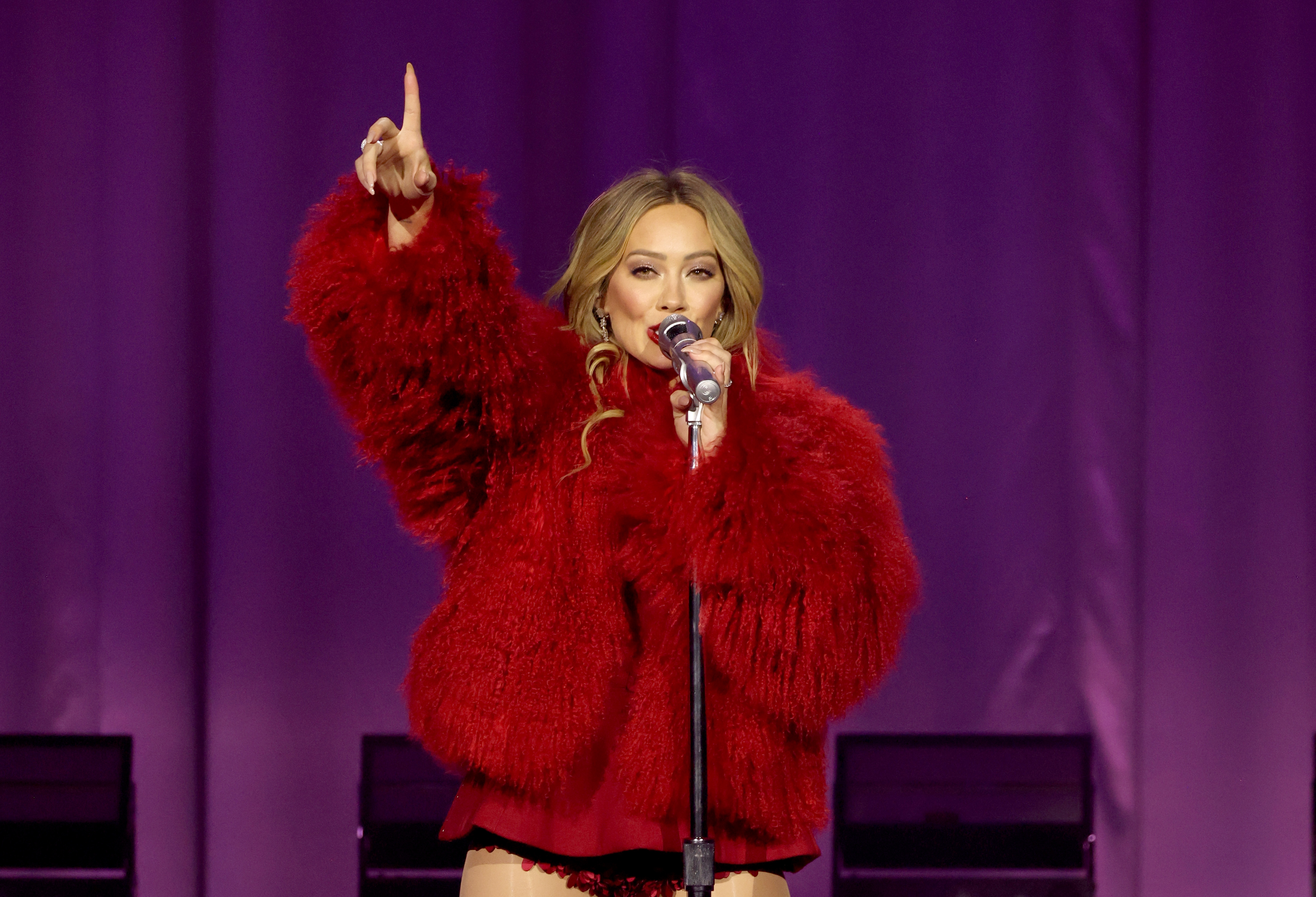 Hilary Duff performing connected  stage, wearing a plush outfit, holding a microphone, and gesturing upwards with 1  hand