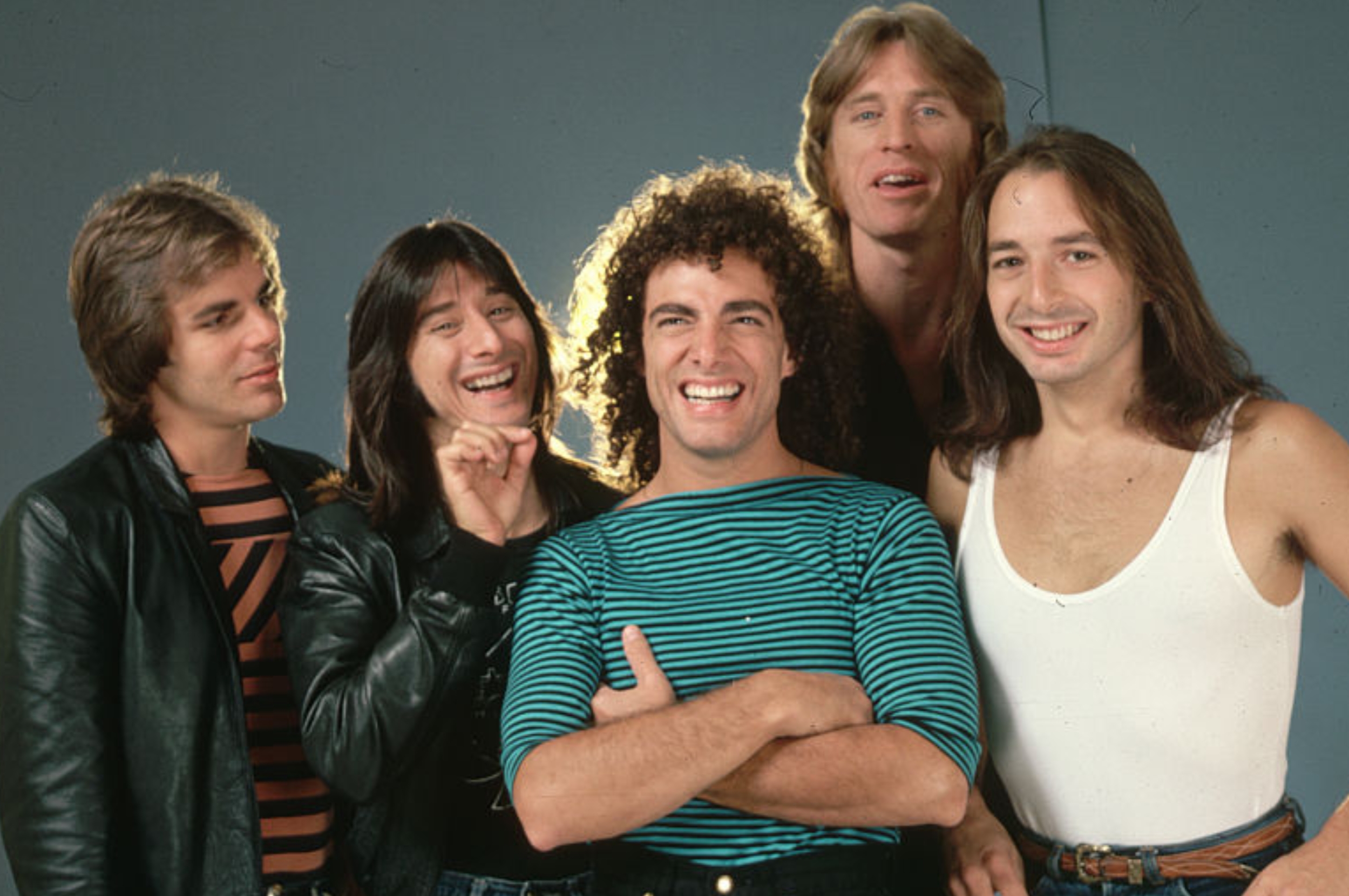 Five people smiling, wearing 1970s-style casual clothing: leather jacket, striped tops, and tank top. Group poses together against a plain backdrop