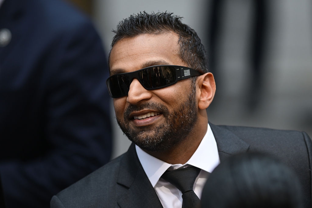 En person i dress og solbriller smiler selvsikkert på et utendørsarrangement