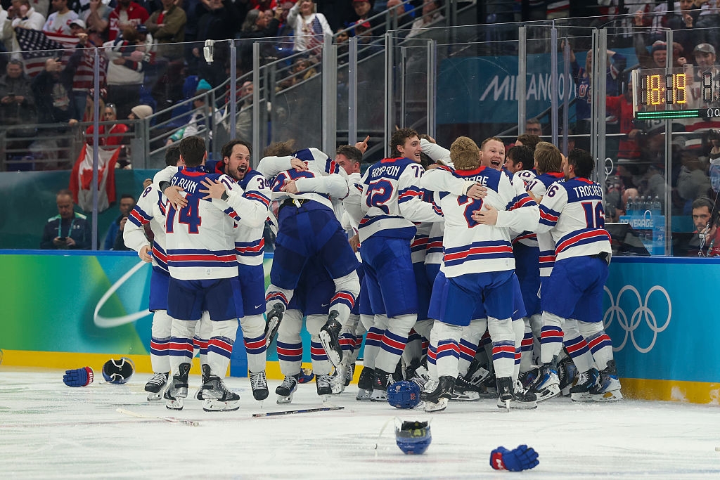 Hockeylag i trøyer feirer på is etter å ha vunnet olympisk kamp, ​​med hjelmer spredt rundt. Folkeklokker i bakgrunnen