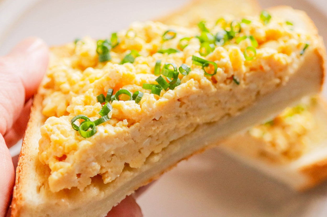 手に持たれた厚切りパンに、スクランブルエッグとネギがのったサンドイッチが見える。