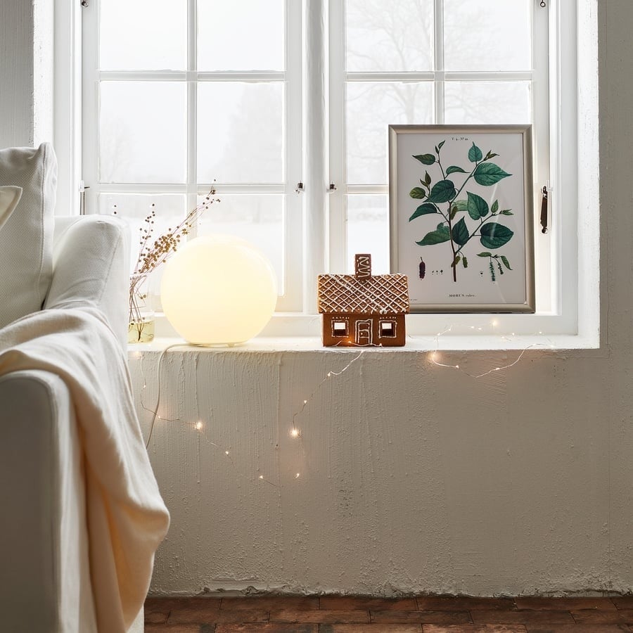 Cozy windowsill with a glowing lamp, framed botanical print, small decorative house, and string lights, creating a warm atmosphere