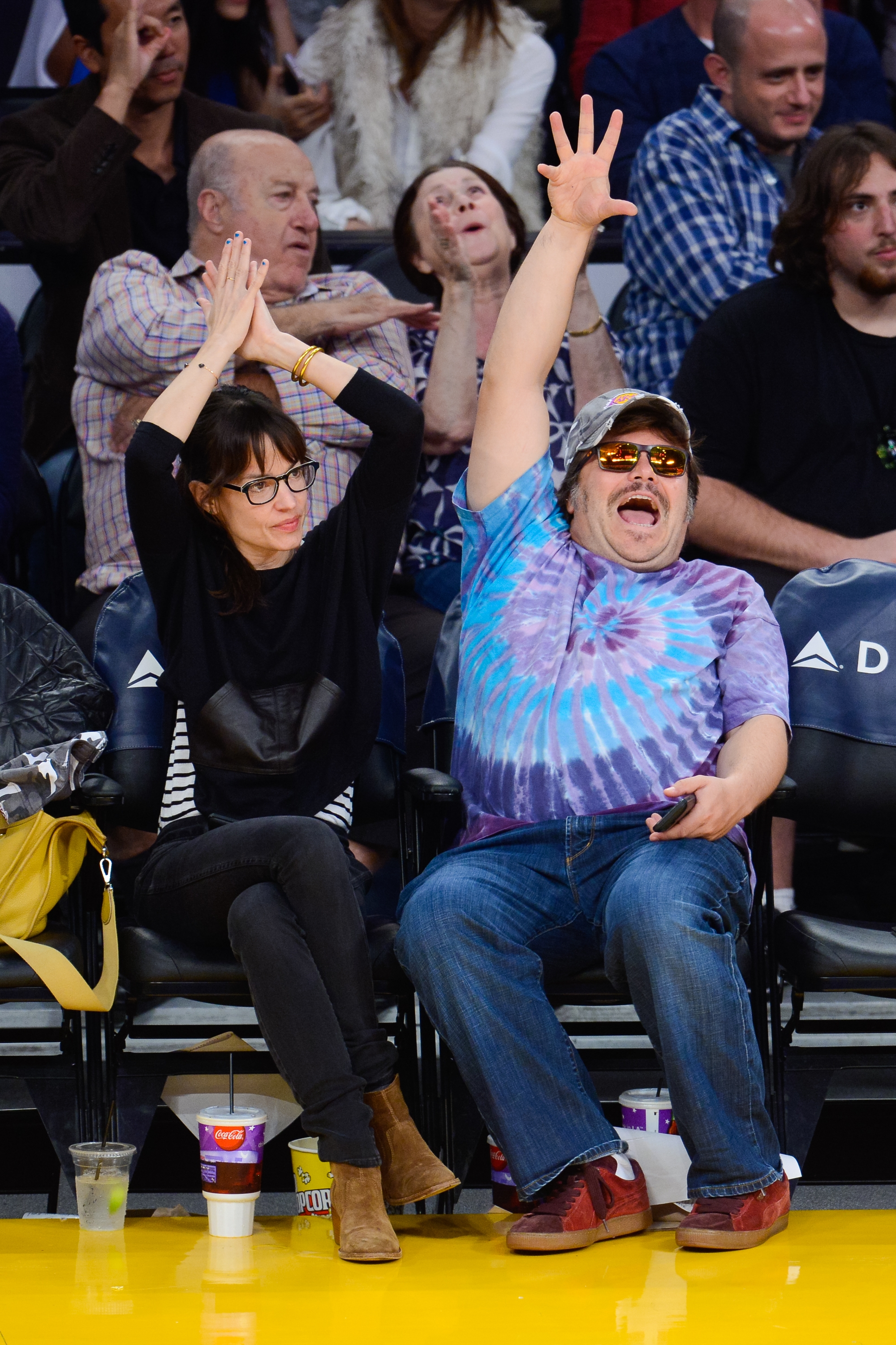 People enthusiastically cheering astatine a sports event; 1 waves successful a tie-dye garment and sunglasses, the different claps successful a achromatic sweater