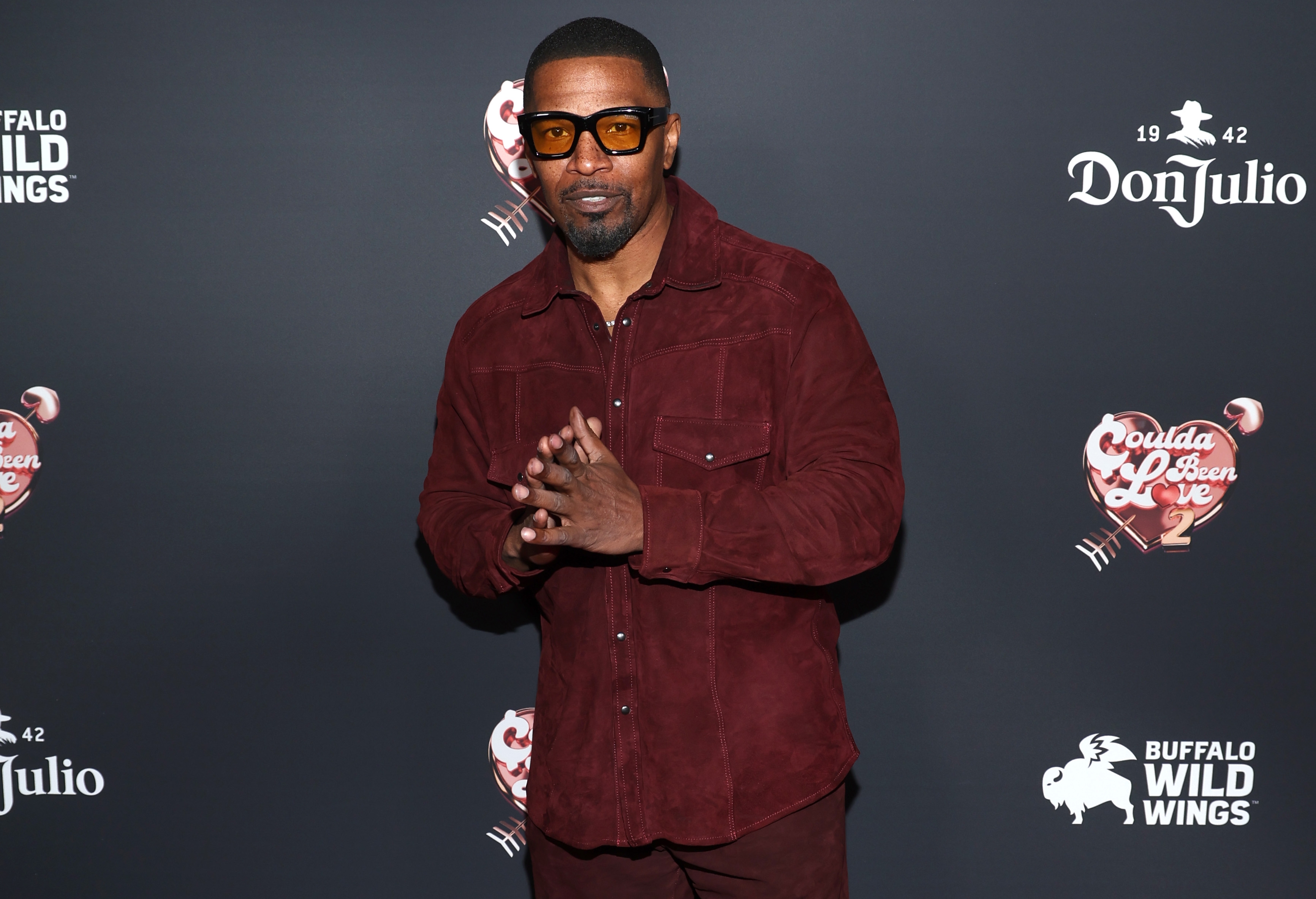 Man connected  reddish  carpet successful  casual overgarment   and sunglasses, lasting  against a backdrop with logos, hands clasped together