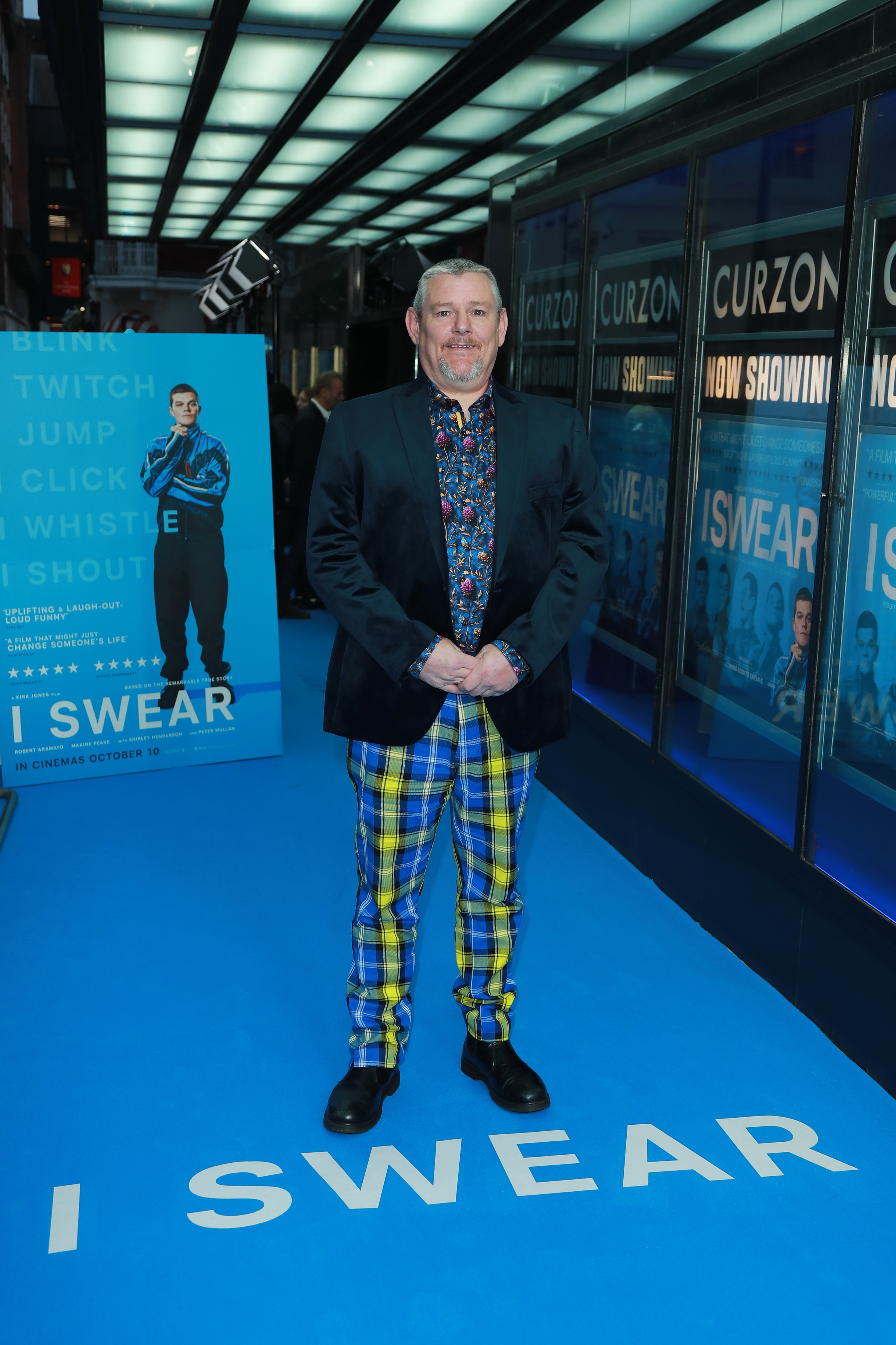 Man successful  patterned garment  and plaid pants astatine  a movie  premiere connected  a bluish  carpet, lasting  successful  beforehand   of movie   posters and marquee