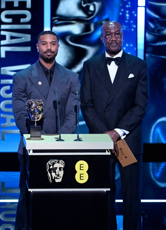 Two men successful  tuxedos basal   connected  signifier    astatine  an awards ceremony, with a trophy connected  the podium betwixt  them